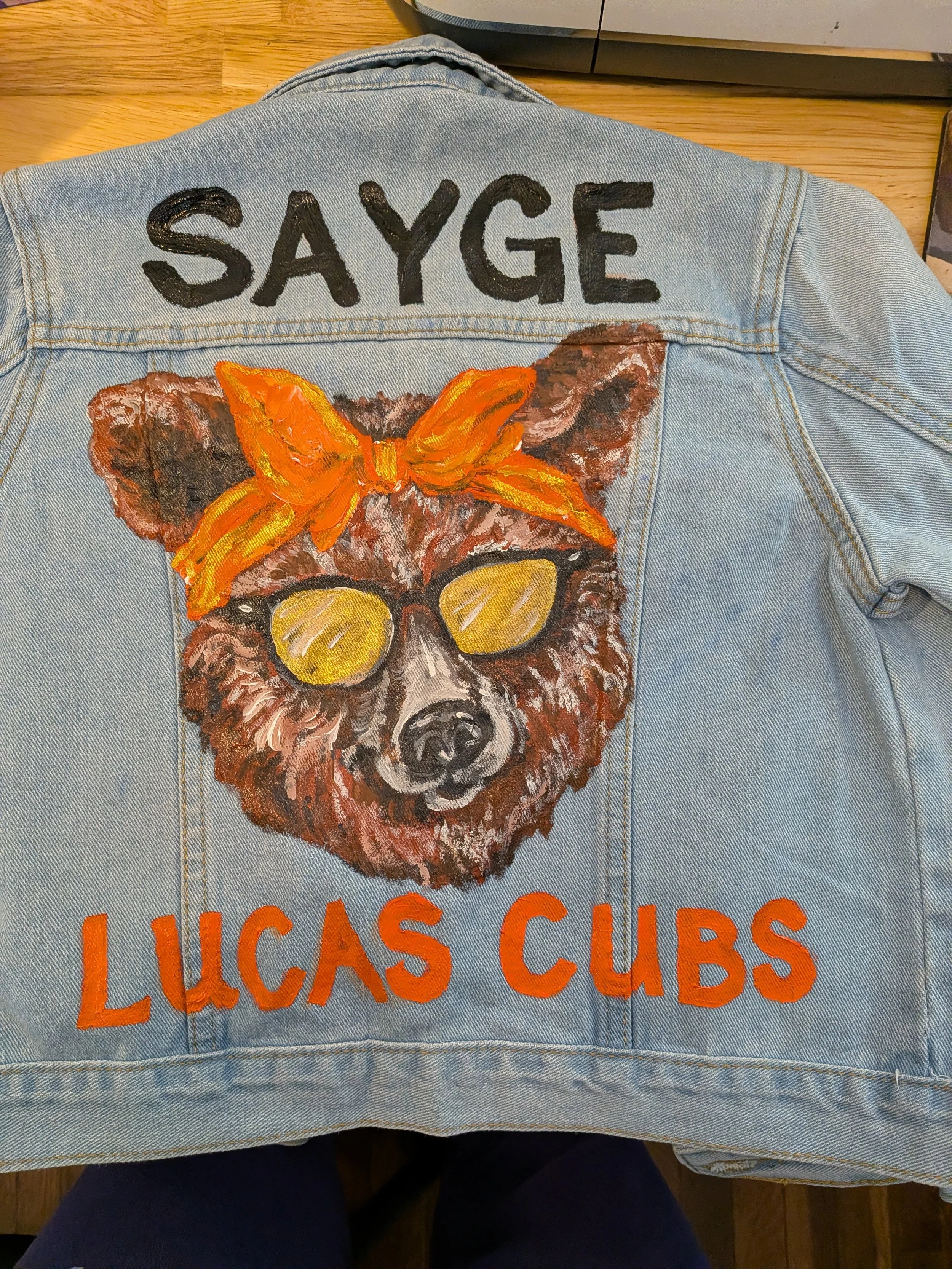 Light blue denim jacket with the word "SAYGE" painted in black on the top part, a large painted bear face wearing sunglasses and an orange bandana in the middle, and the words "LUCAS CUBS" painted in orange on the bottom part.