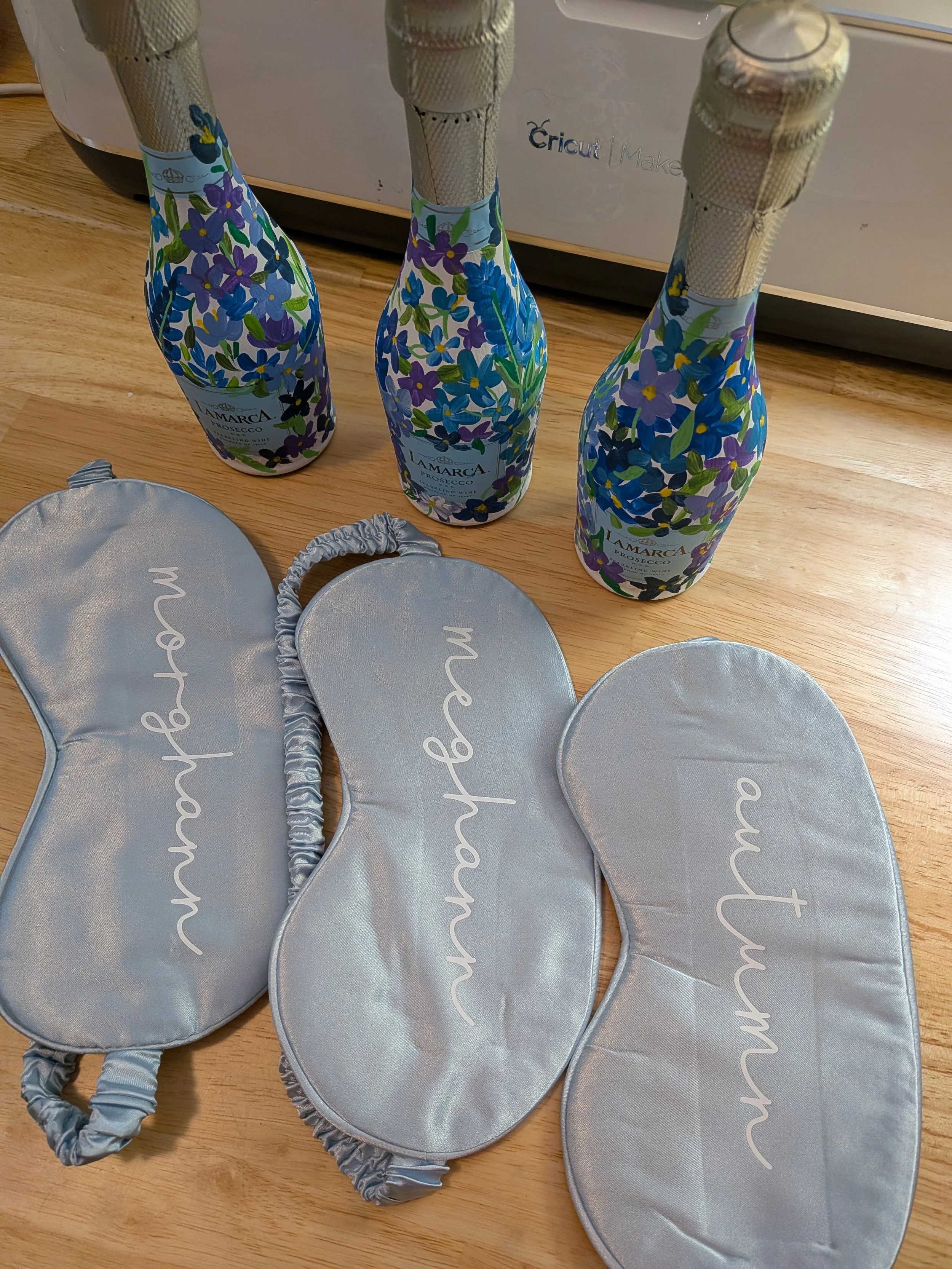 Three bottles of LaMarca Prosecco on a wooden surface in front of a silver Cricut machine. There are also four silver party hats with the word 'happy' written on them, placed on the wooden surface in front of the bottles.