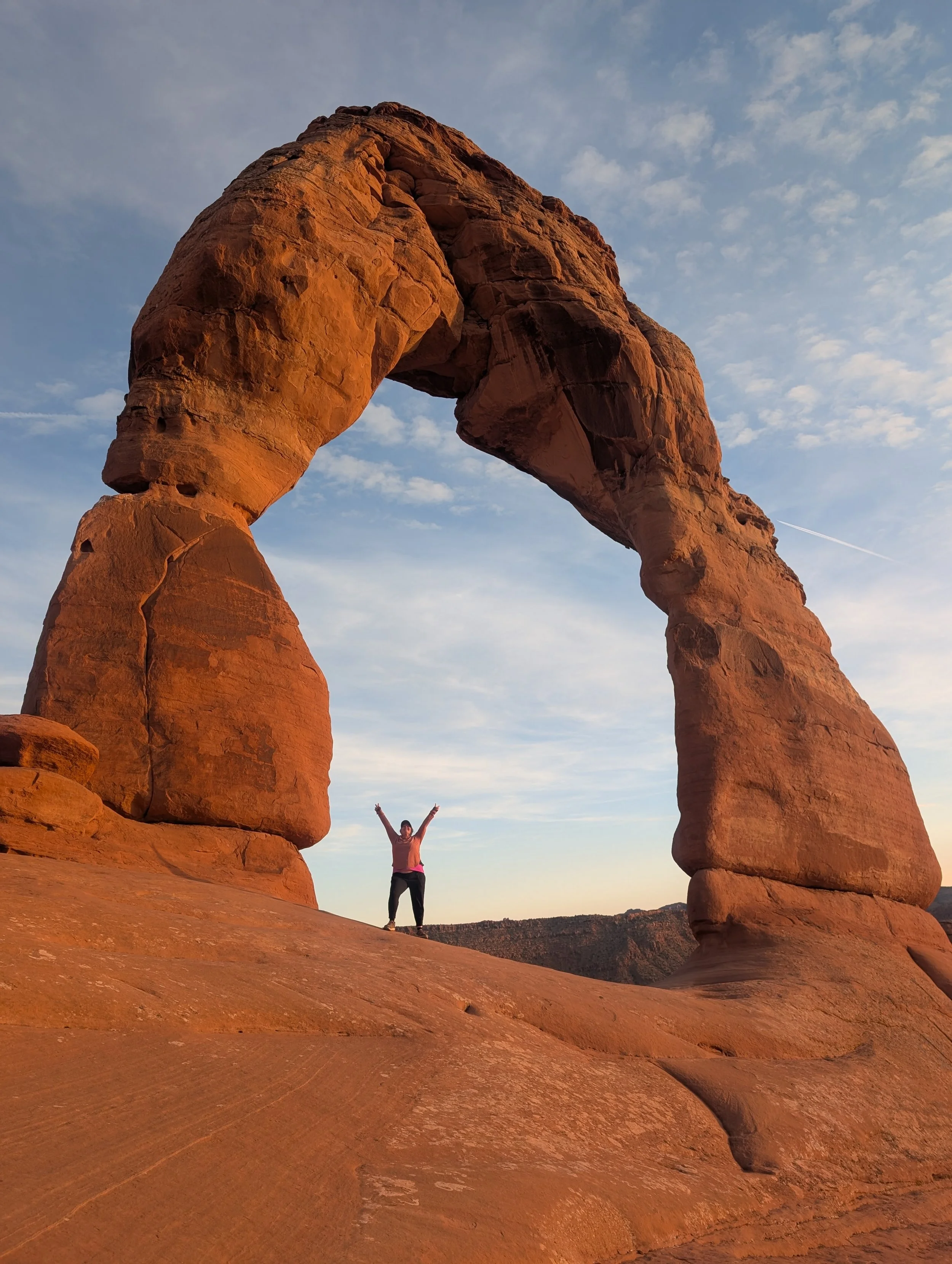 The artist with her arms raised standing beneath a large natural rock formation resembling a horseshoe arch during sunset.