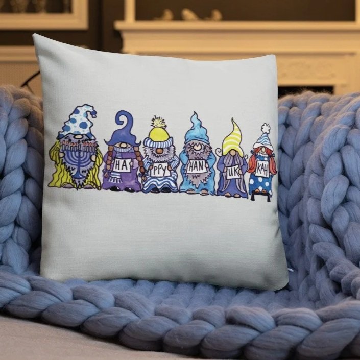 Decorative pillow with cartoon gnomes wearing colorful hats, holding cards that spell out "HAPPY HANUKKAH," sitting on a chunky knit blanket.