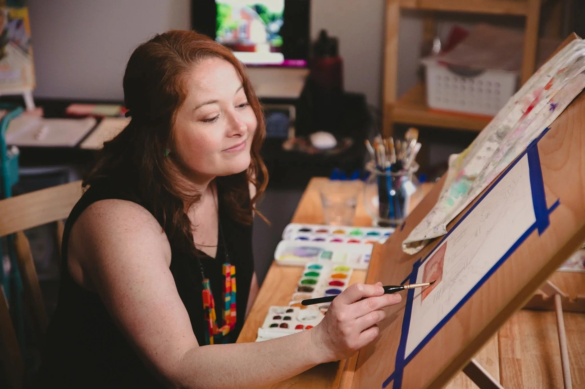 A woman with auburn hair and colorful earrings is painting on a canvas held on an easel. She is seated at a wooden table with watercolor paints, brushes, and a palette. The background shows shelves with art supplies and a TV.