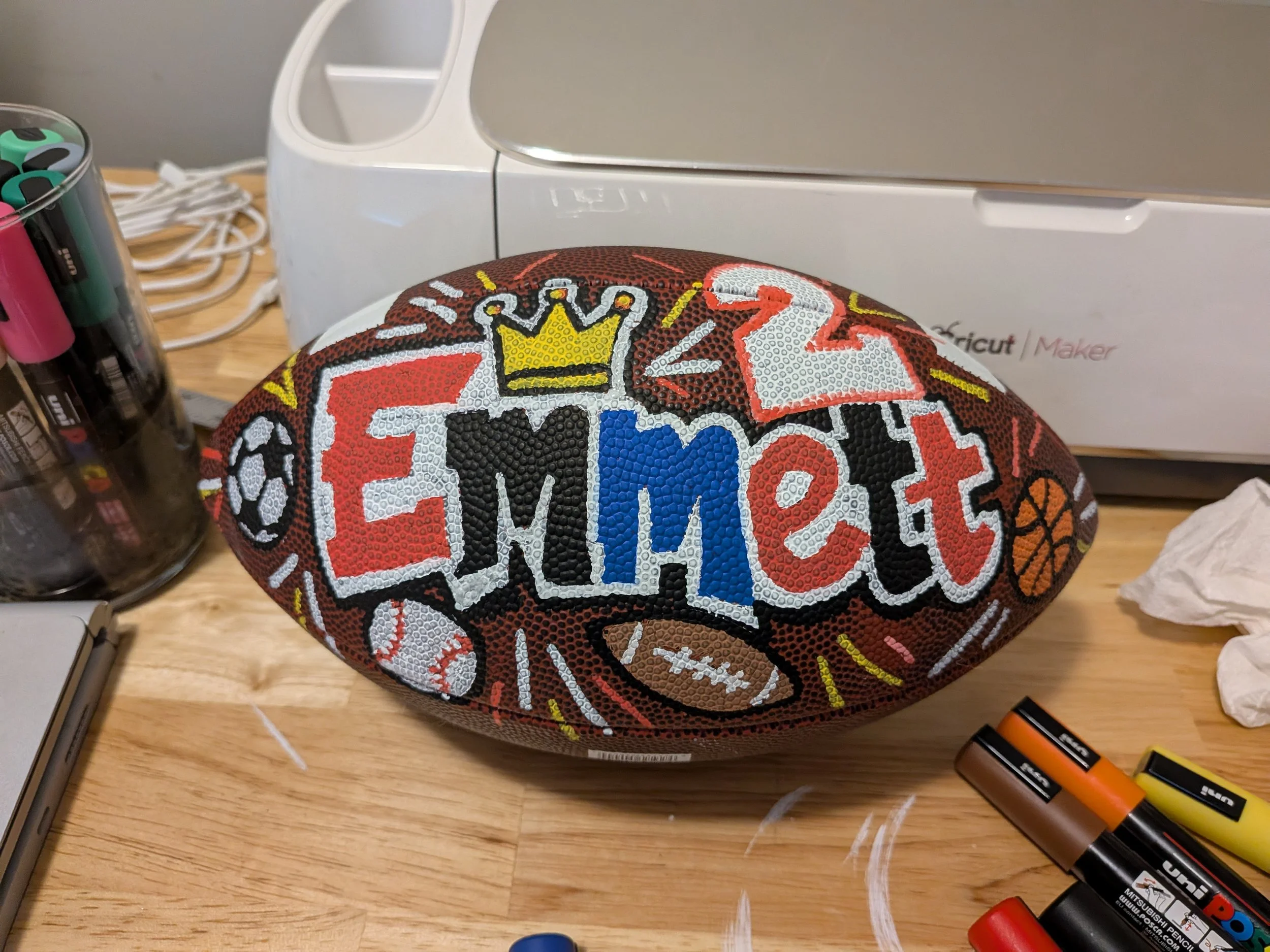 Close-up of a textured basketball with a colorful design and the words 'Team Up' on it, on a wooden table surrounded by markers, batteries, and miscellaneous objects.