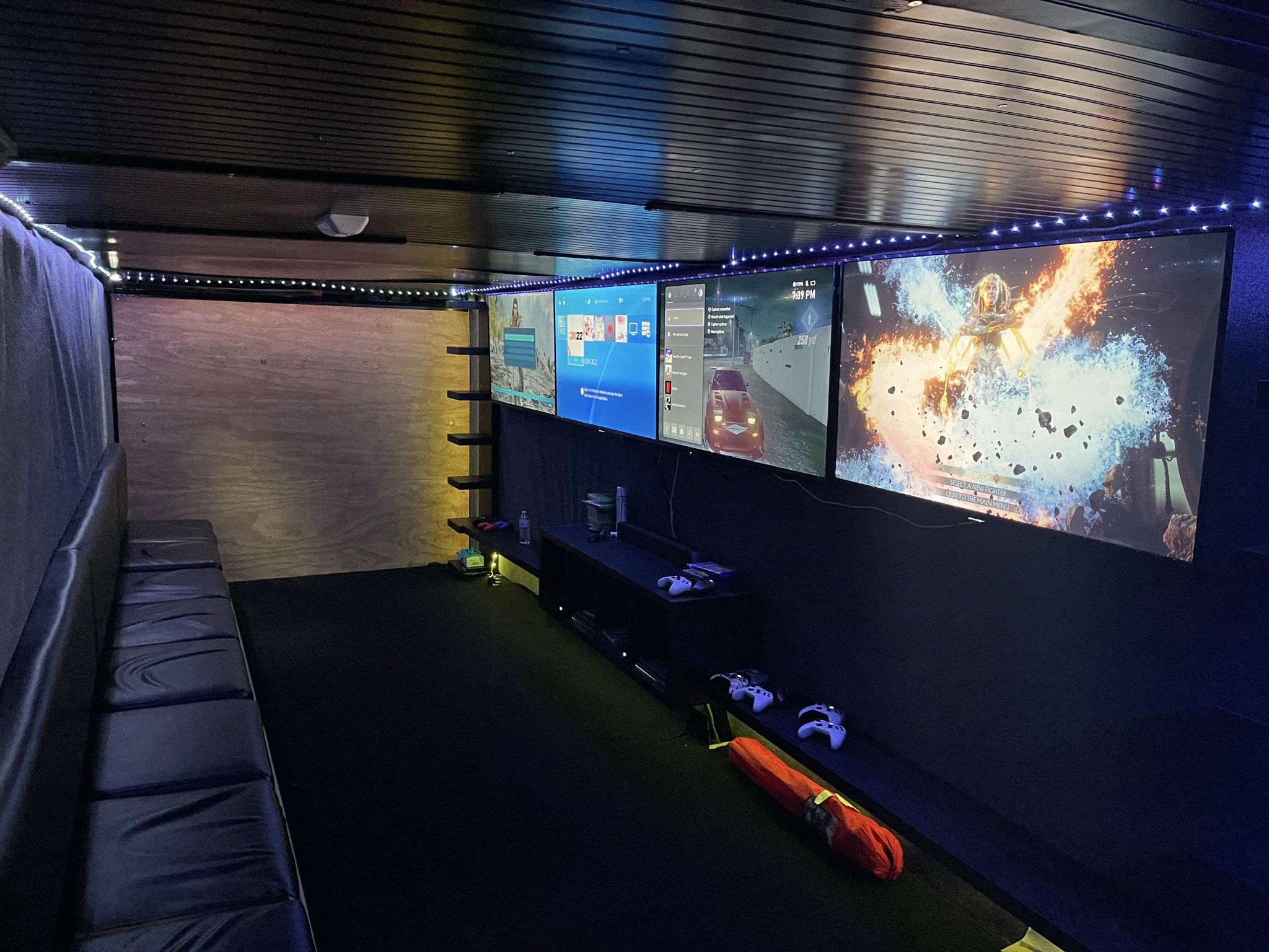 A gaming room with three large screens on the wall, showing different digital displays, including a game with an explosion scene; black leather seating along the left side, and gaming controllers on a shelf and table. The ceiling has wood paneling an