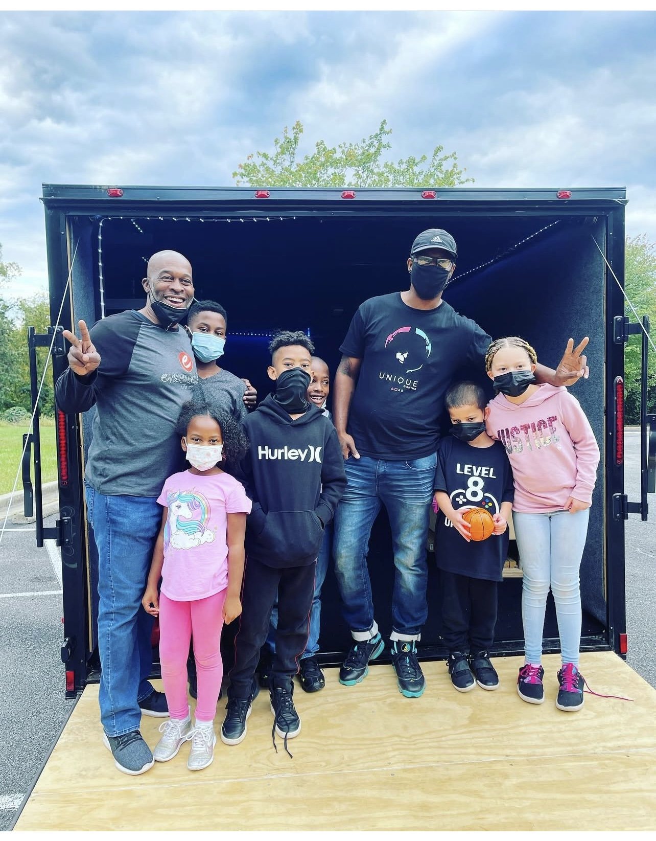Group photo of children and two adults, standing in front of a game truck with a wooden platform at a park, during a birthday celebration