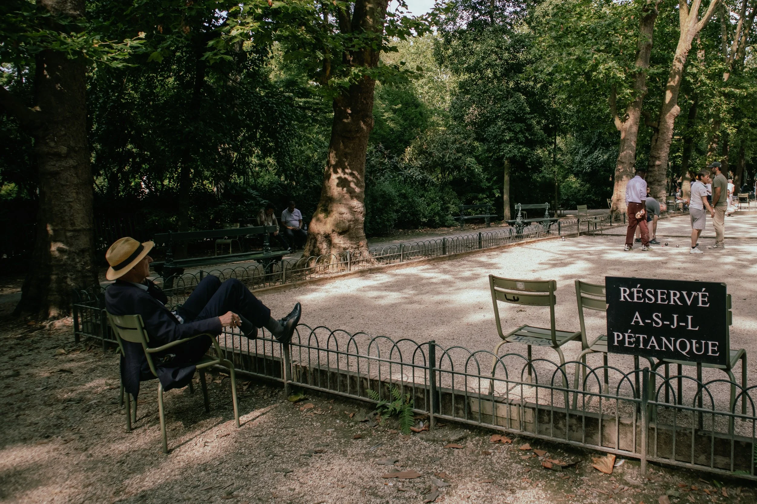 A man wearing a hat and sitting on a chair in a park, with a sign indicating the pétanque court is reserved. Several people are playing pétanque in the background among tall trees.