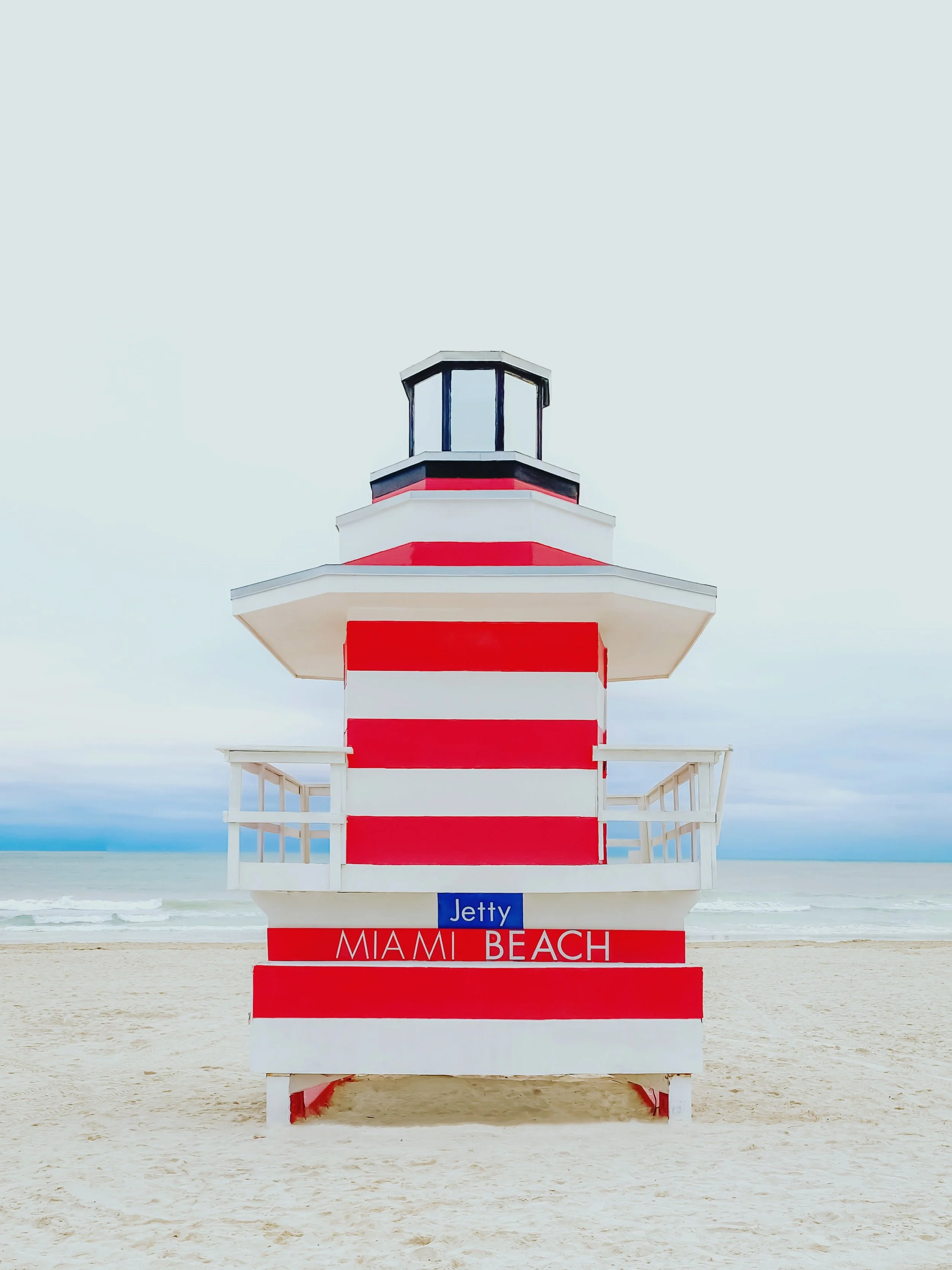 A red and white striped lifeguard tower on a sandy beach near the ocean with a cloudy sky.