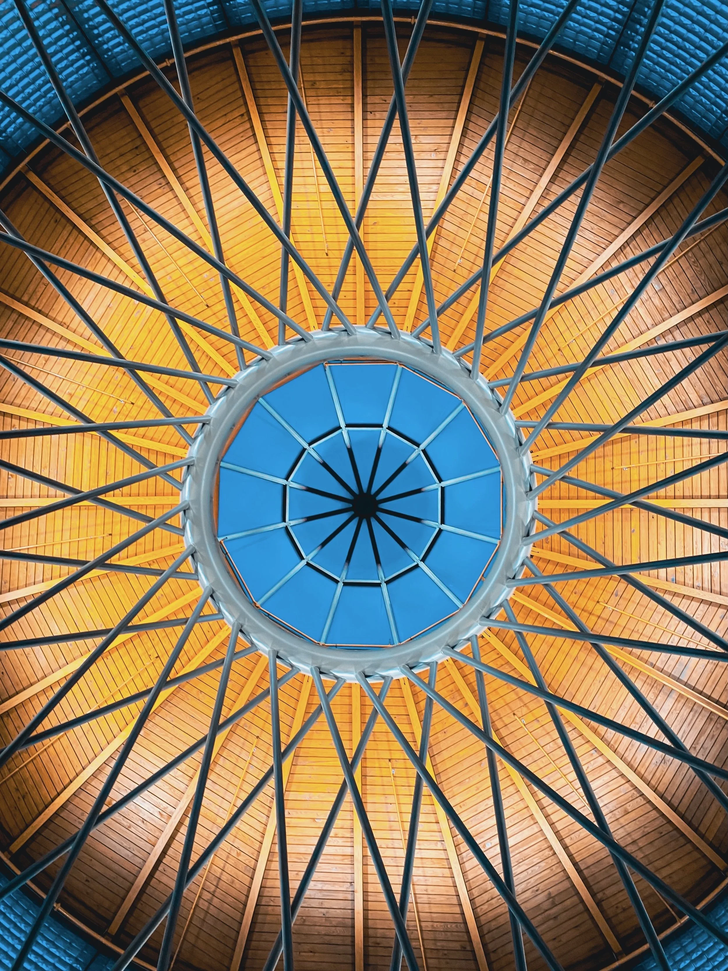 Looking up at the interior of a dome-shaped structure with a central circular opening, wooden panels on the ceiling, and metal spokes radiating outward.