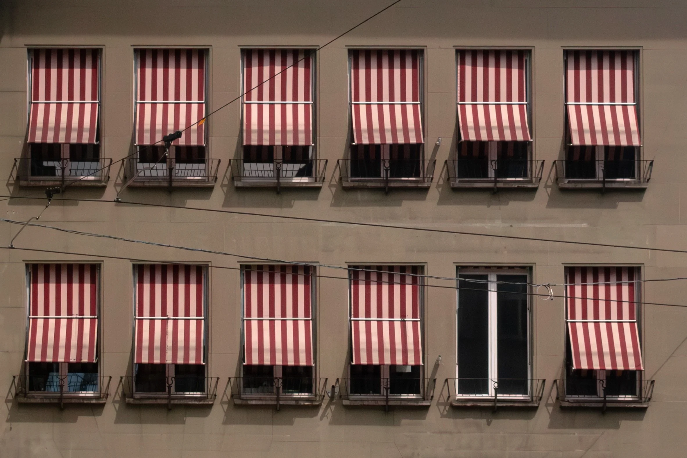 Facade of a building with three floors, each with four windows fitted with red-and-white striped awnings and small balconies, with wires crisscrossing in front.