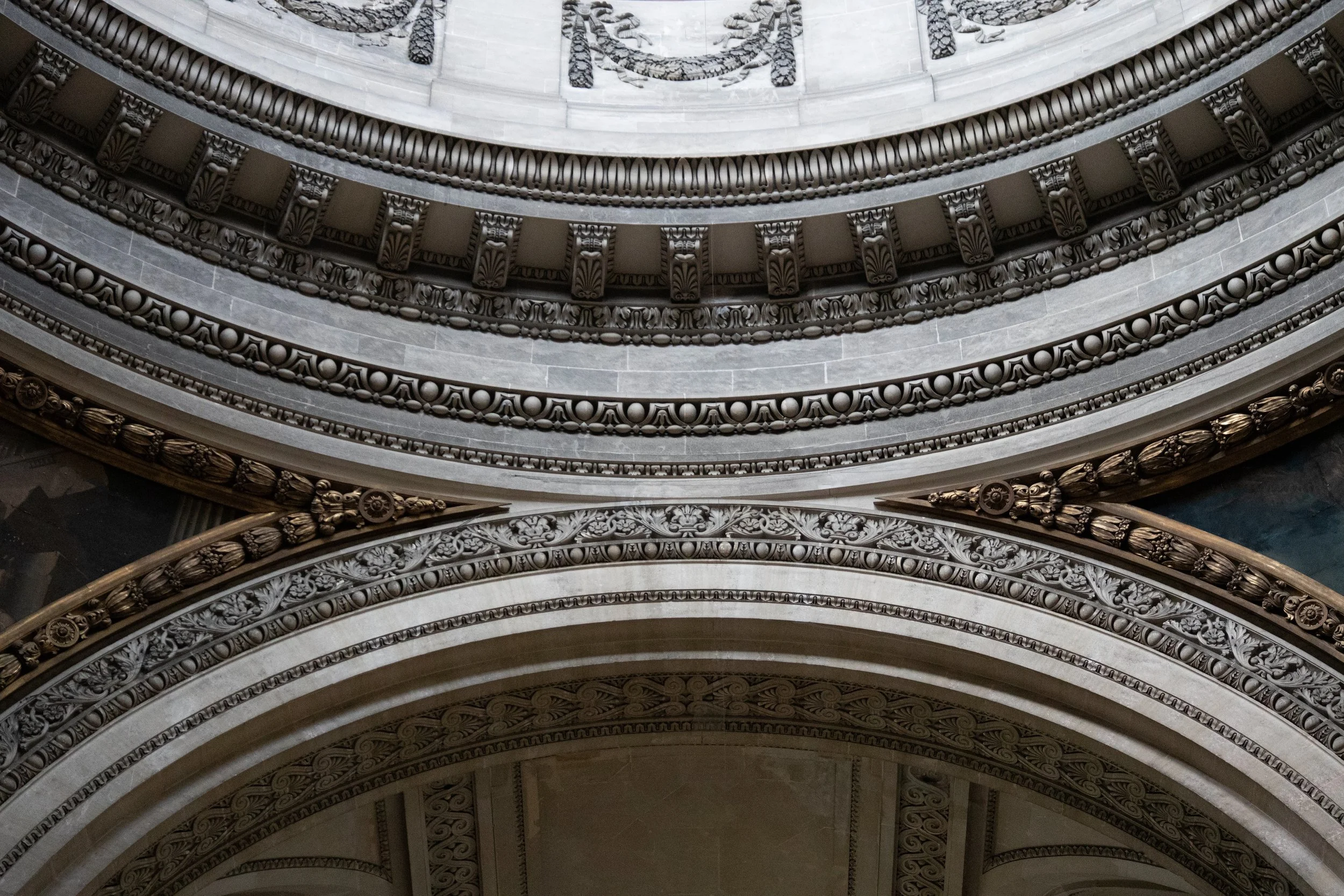 Close-up view of ornate architectural details on a domed ceiling, featuring decorative moldings, intricate carvings, and scalloped designs in shades of gray and gold.