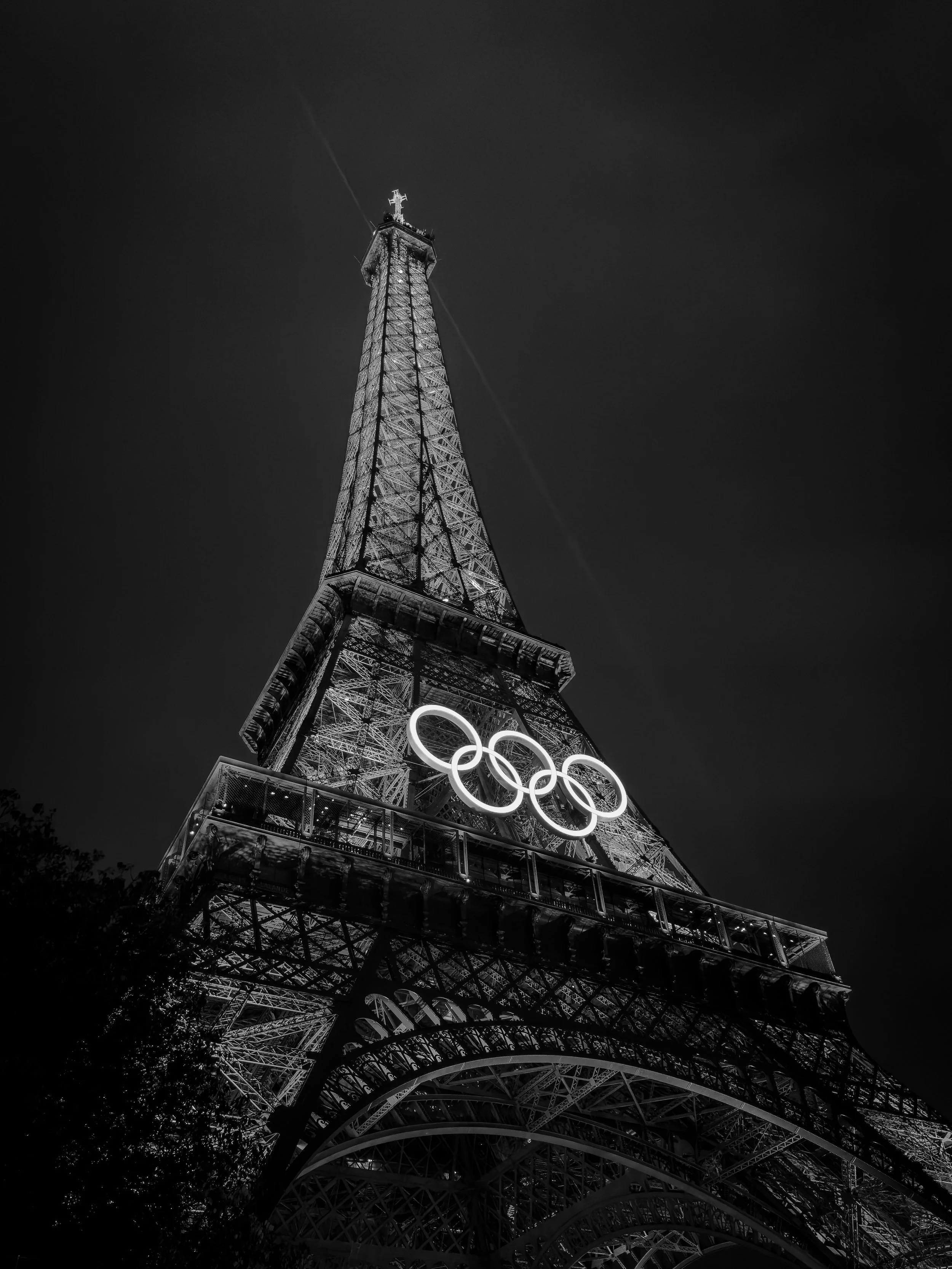 Nighttime black and white photo of the Eiffel Tower in Paris, with the Olympic rings illuminated on the lower part of the tower.