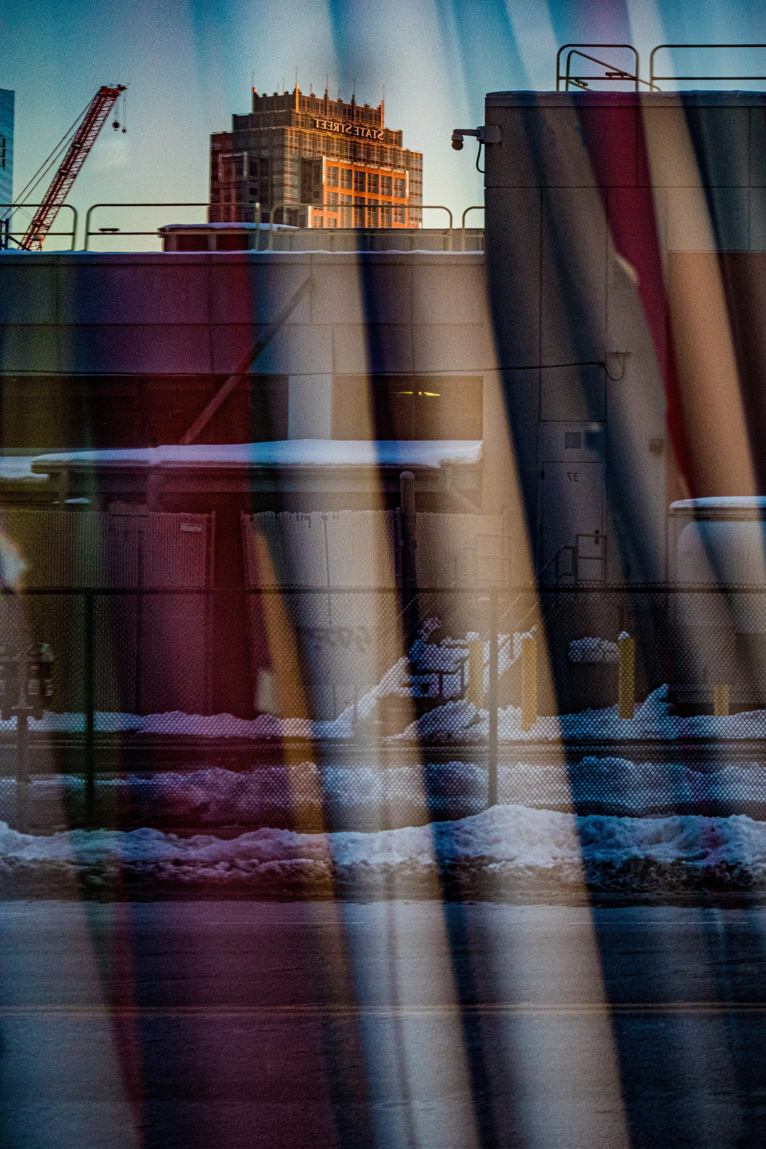 Reflection of a cityscape with a building labeled 'State Street' and construction crane, seen through window blinds, with snow on the ground during late afternoon or sunset.