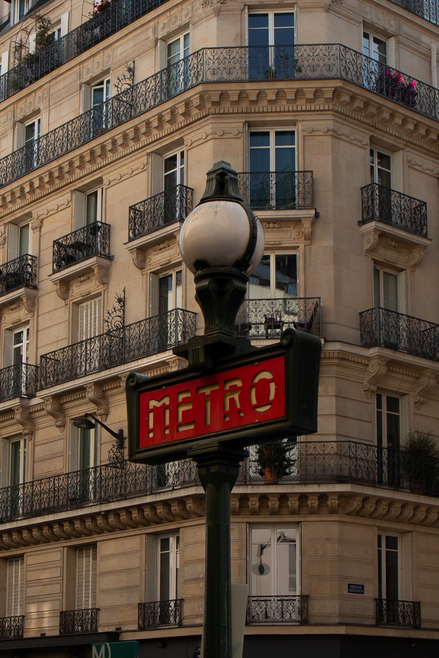 An ornate Paris metro sign with red letters on a black background, mounted on a lamp post in front of a classic Parisian building with decorative wrought iron balconies.