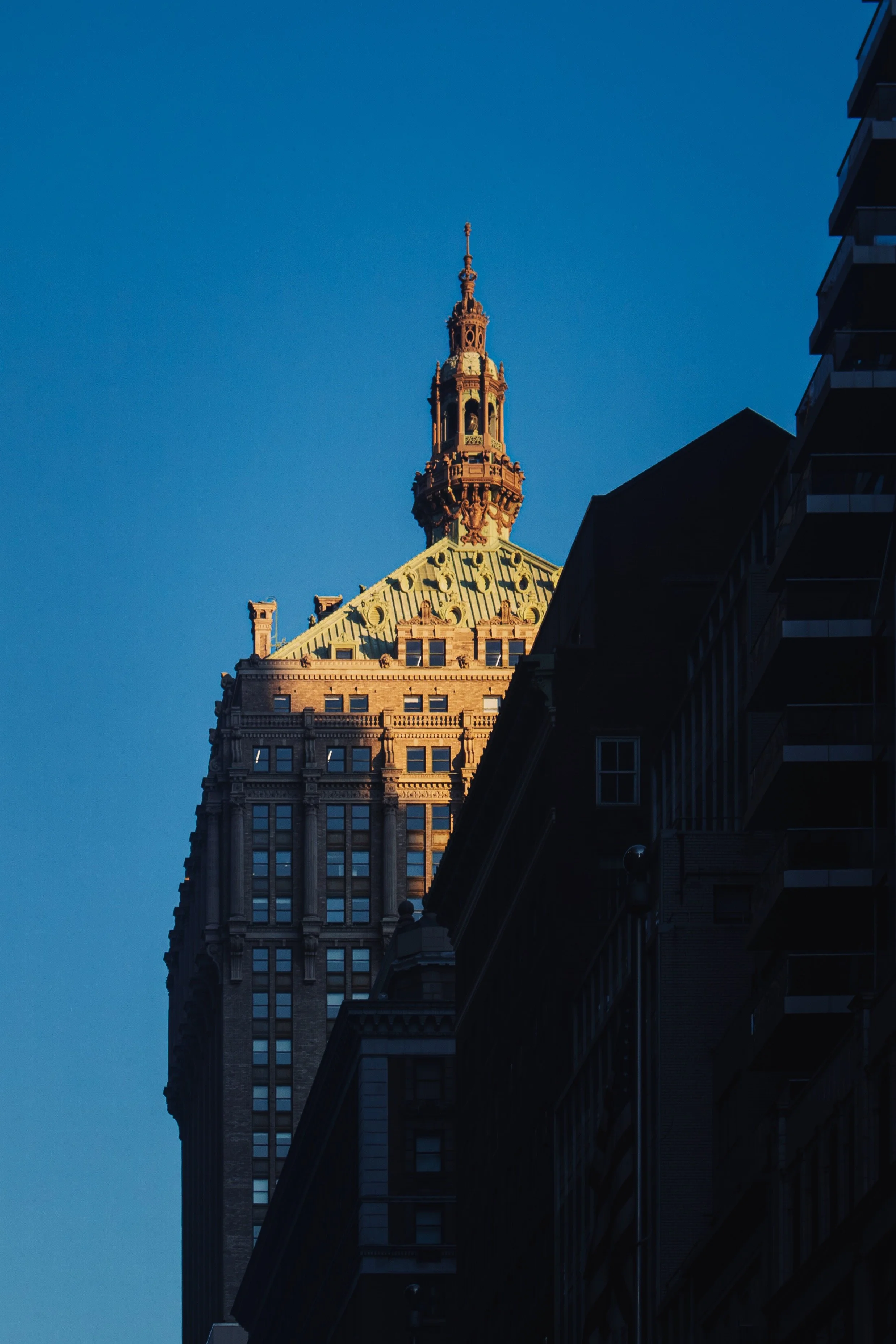 Tall historic skyscraper with ornate architecture, topped with a spire, set against a clear blue sky, with dark shadows from nearby buildings.