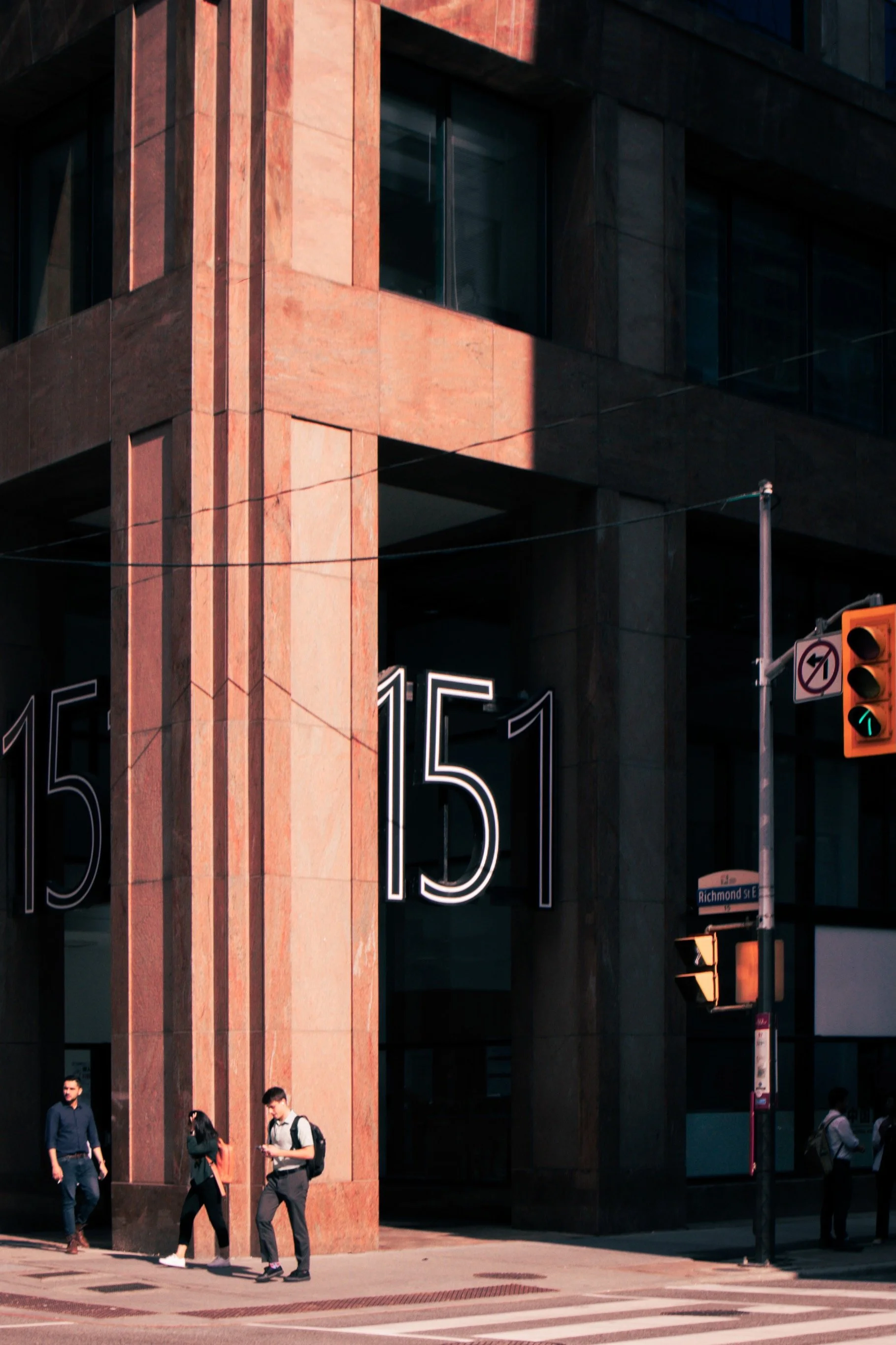 People walking on a city sidewalk in front of a large building with neon signs displaying '151'. A traffic light shows green, and there is a street sign reading 'Richmond St E'.