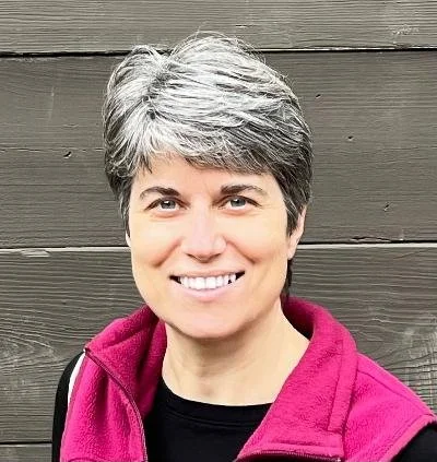 Woman with short gray hair smiling, wearing a black shirt and a pink jacket, standing in front of a wooden wall.