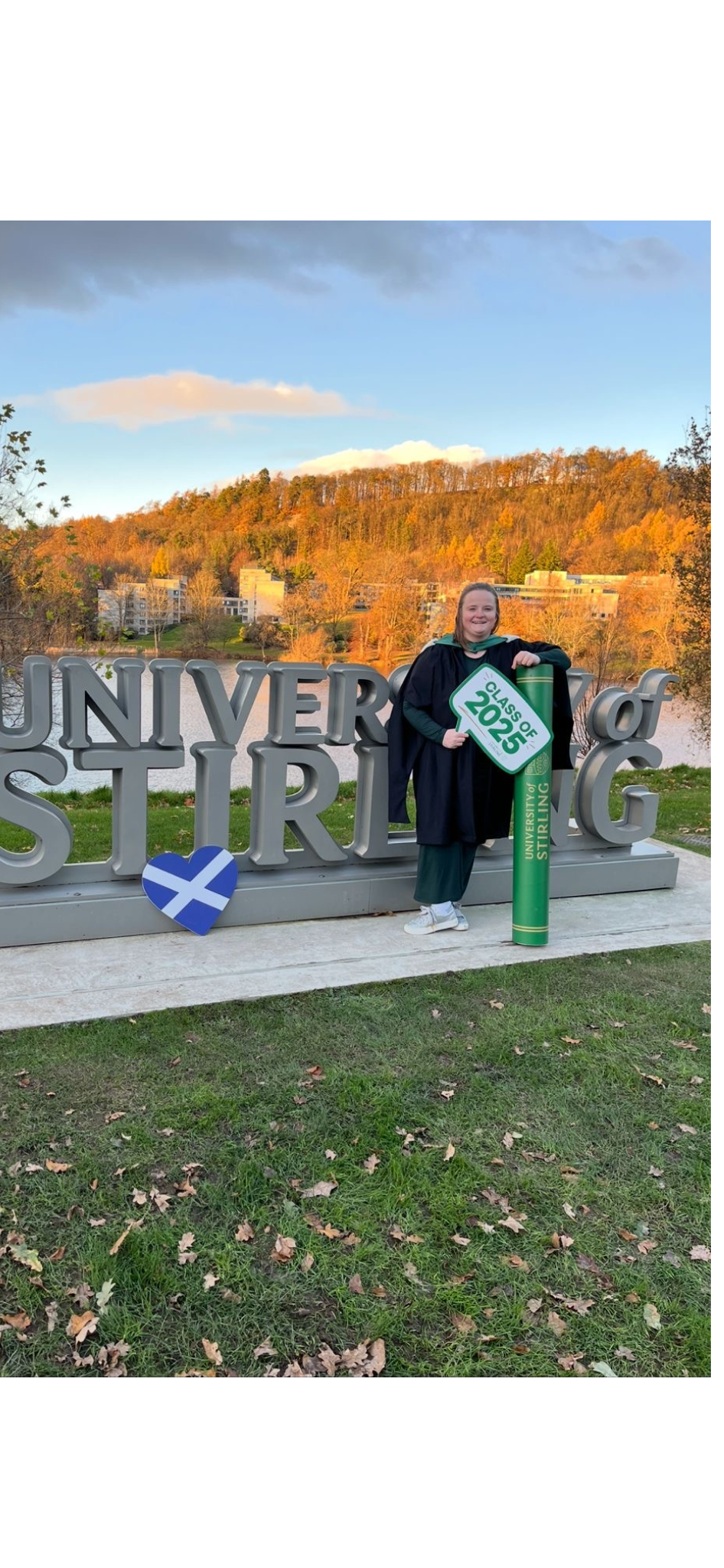 Hope Murray standing in front of the University of Stirling sign, holding a Class of 2025 marker and graduation scroll, with autumn trees and campus buildings in the background.