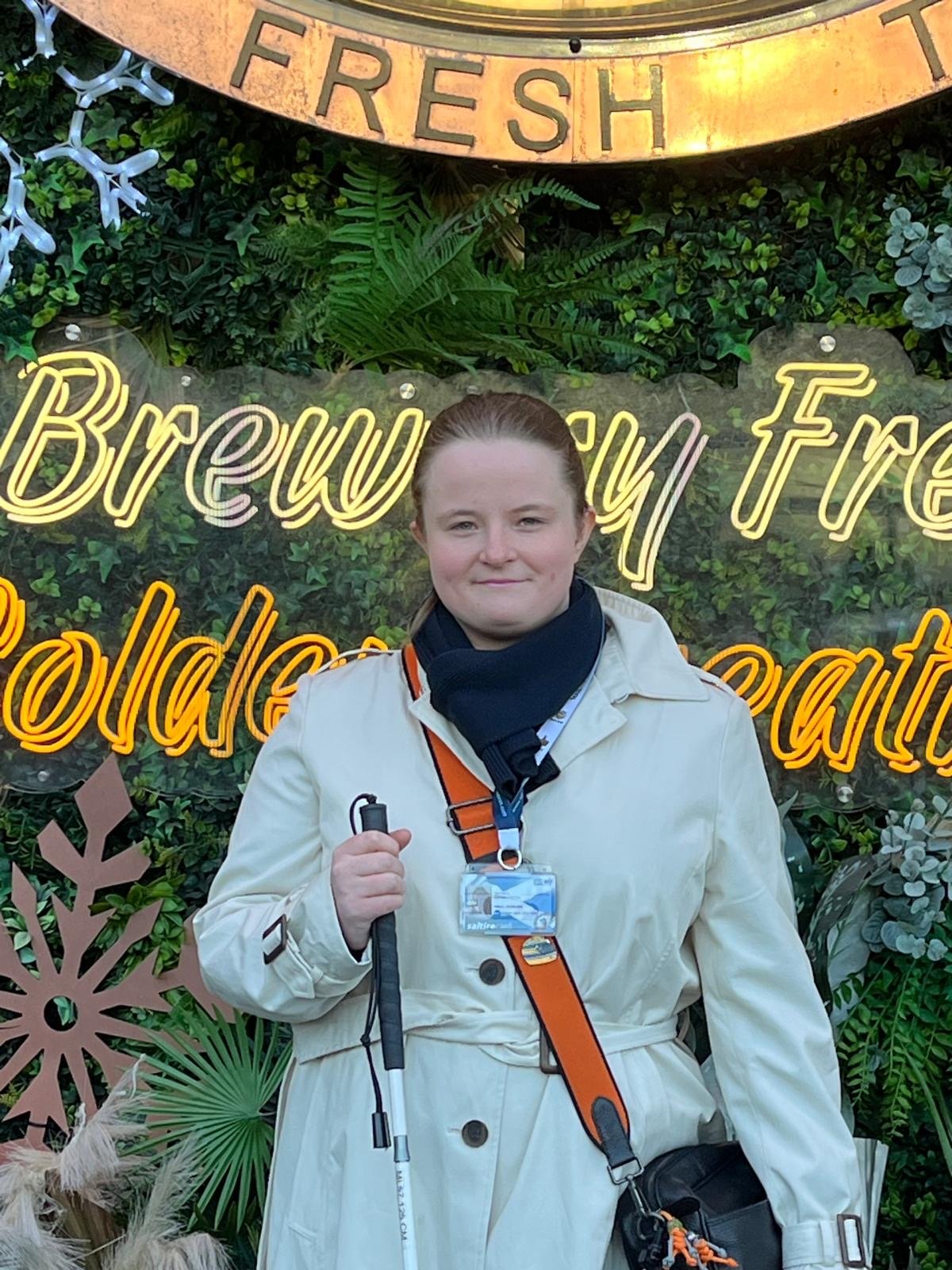 Hope standing outdoors holding a long cane, in front of illuminated lettering and green foliage decorations.