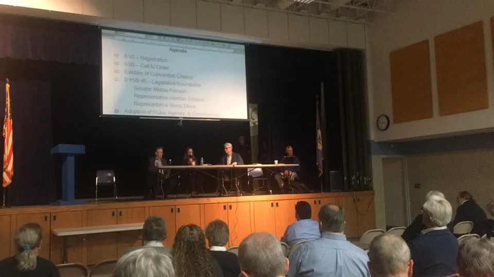 Photo of a school auditorium. On the stage are four people who are leading a convention. On the ground level are people seated, their backs to the photographer
