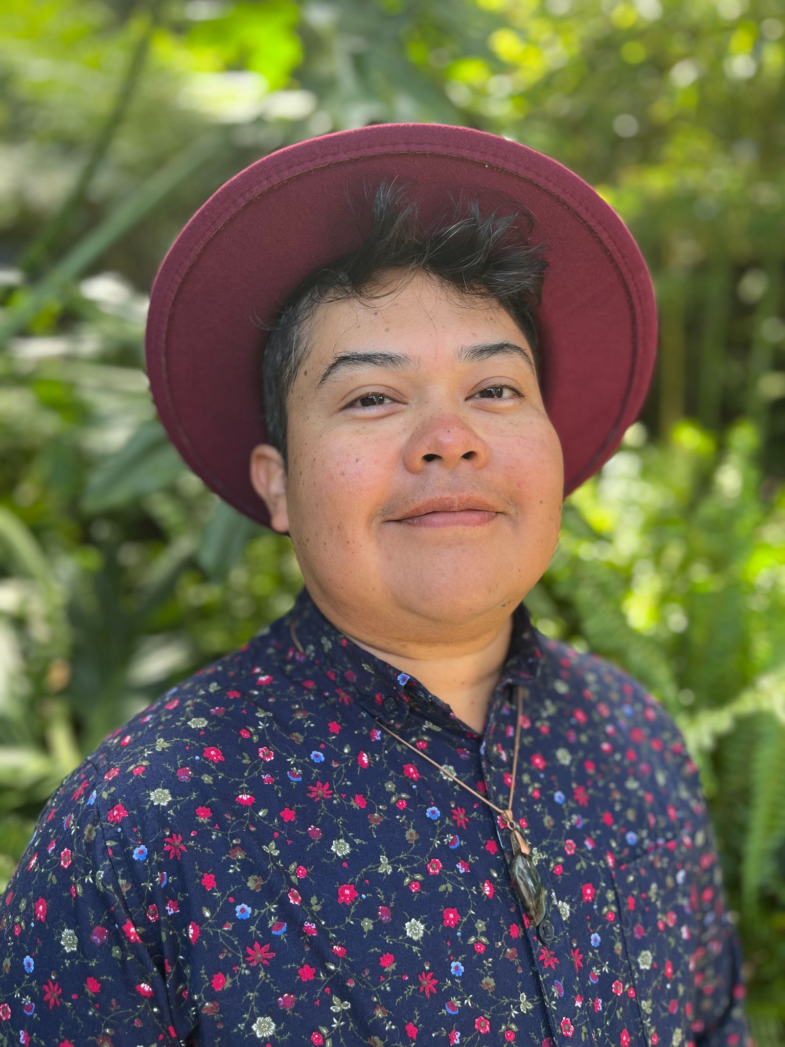 A person with short dark hair wearing a wide-brimmed maroon hat and a navy shirt with a colorful floral pattern, standing outdoors in a garden with green foliage in the background.