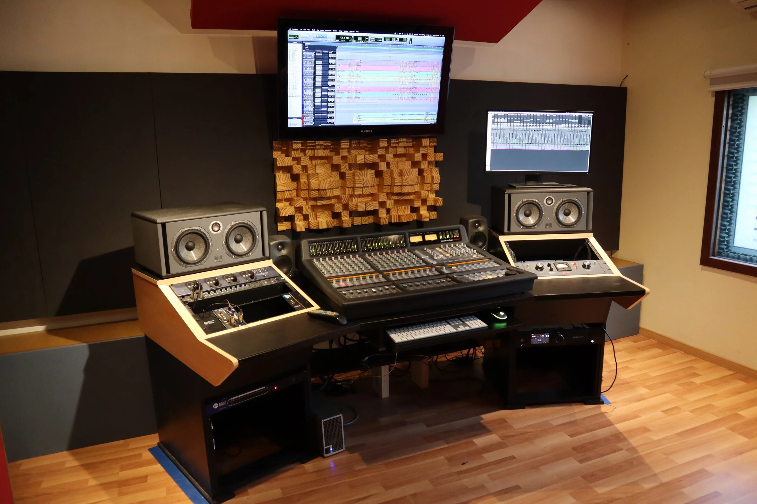 Estudio de grabación con consola de audio, altavoces y monitores de computadora, en una habitación moderna con madera clara y paredes gris oscuro.