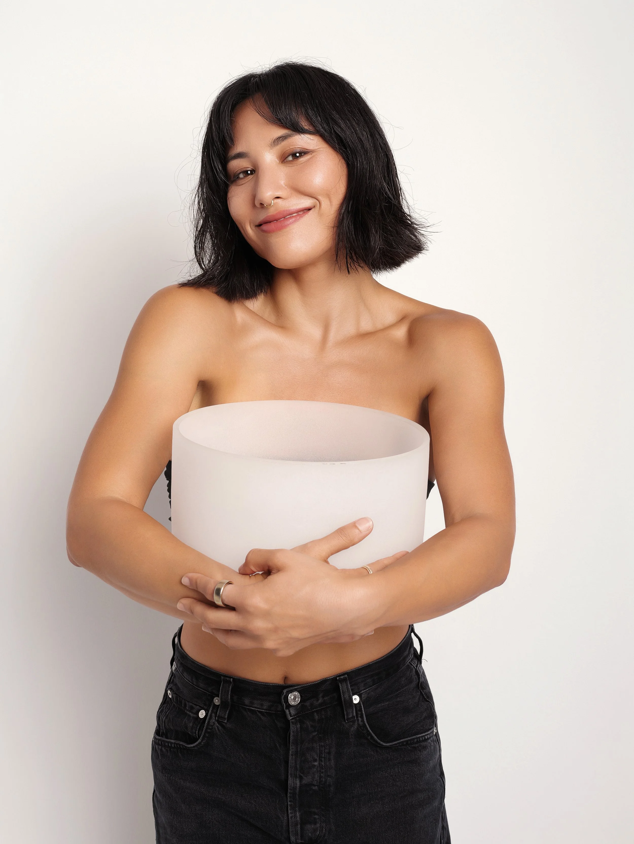 A woman with short dark hair and a septum piercing smiling while holding a white cylindrical object against a white background.