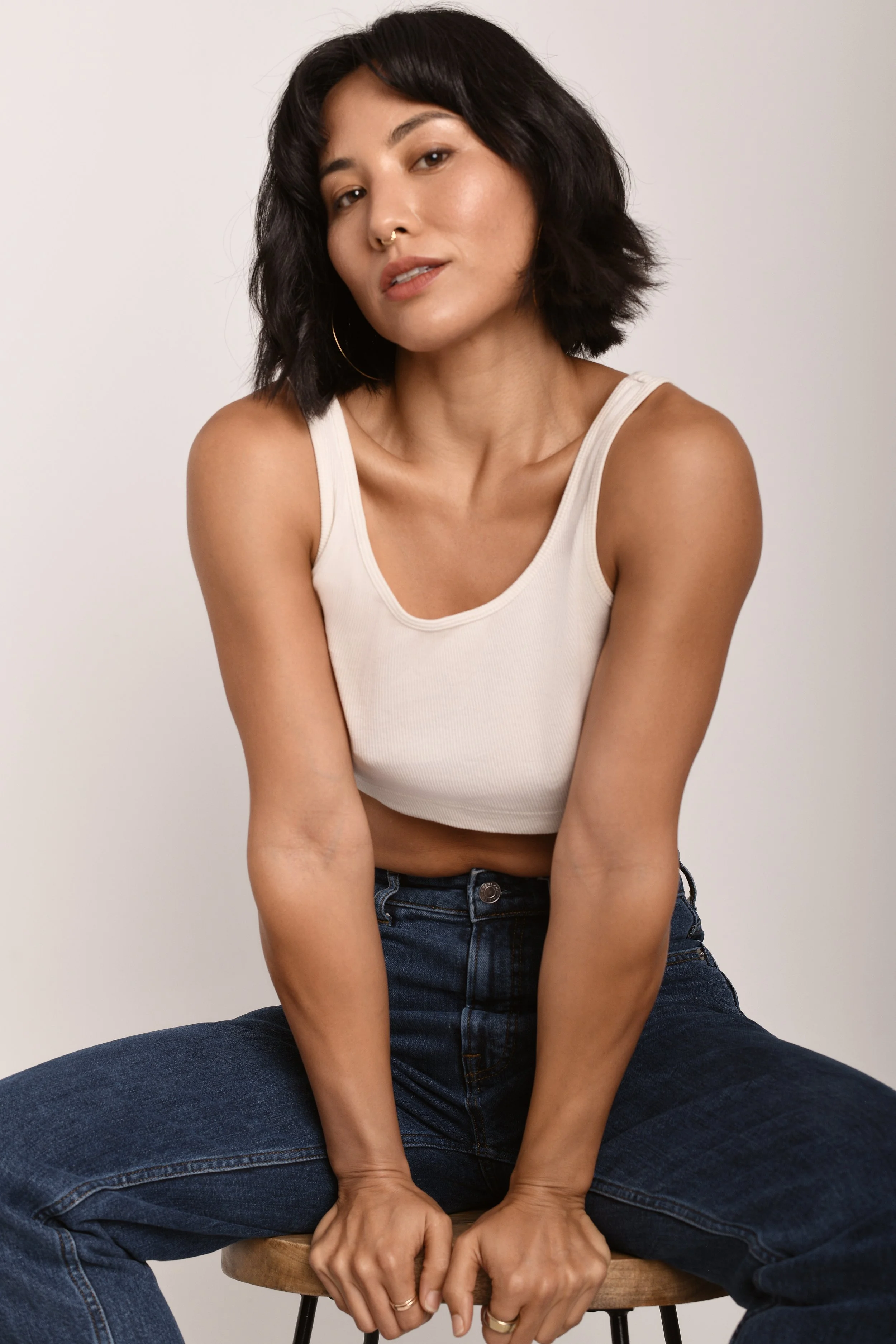 A woman with chin-length black hair, wearing a cream tank top and dark jeans, sitting on a wooden stool against a plain, light-colored background.