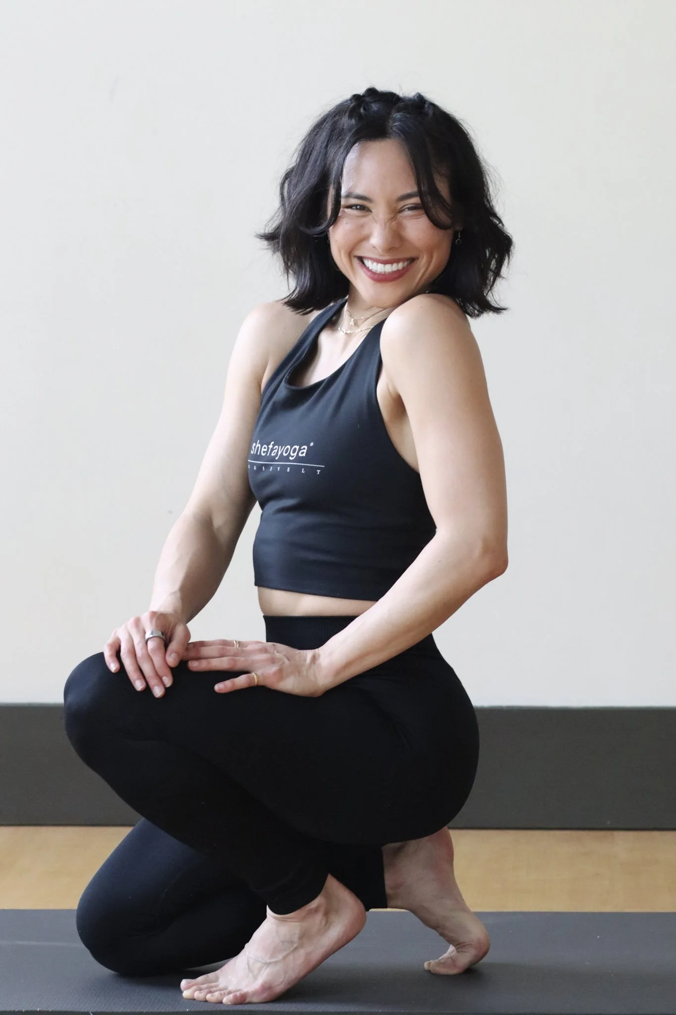 A woman with short dark hair and a black yoga top smiling in a yoga pose on a black mat against a plain white wall.