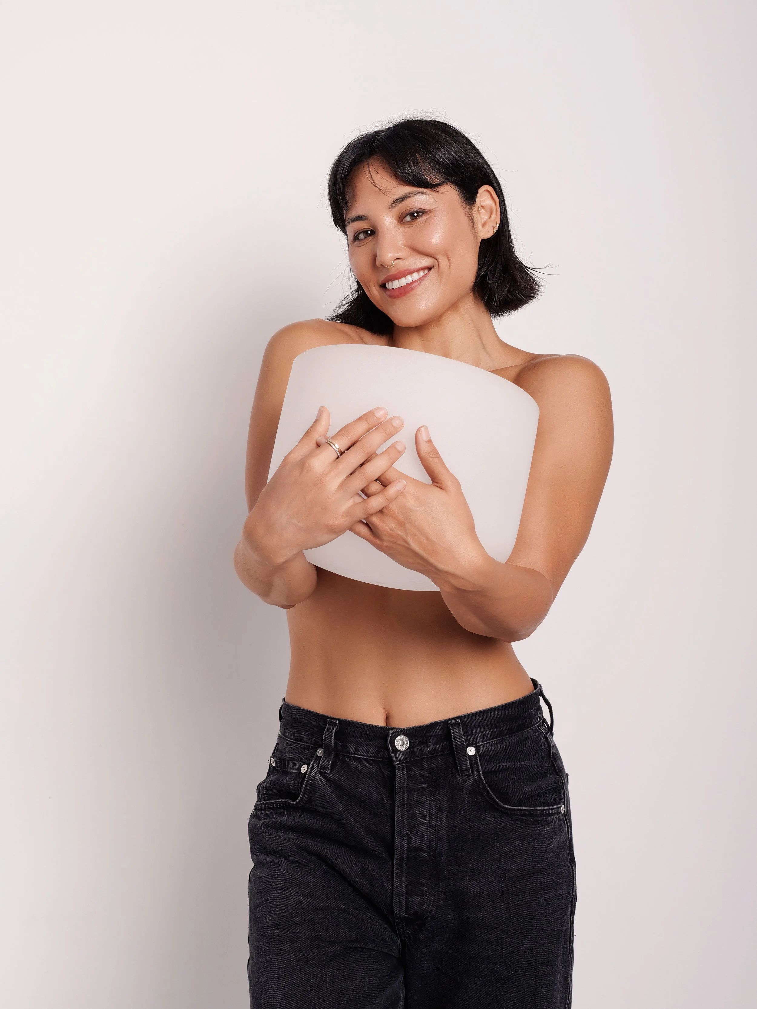A woman with short black hair, smiling and hugging a large white pillow against her chest, standing against a plain white background.