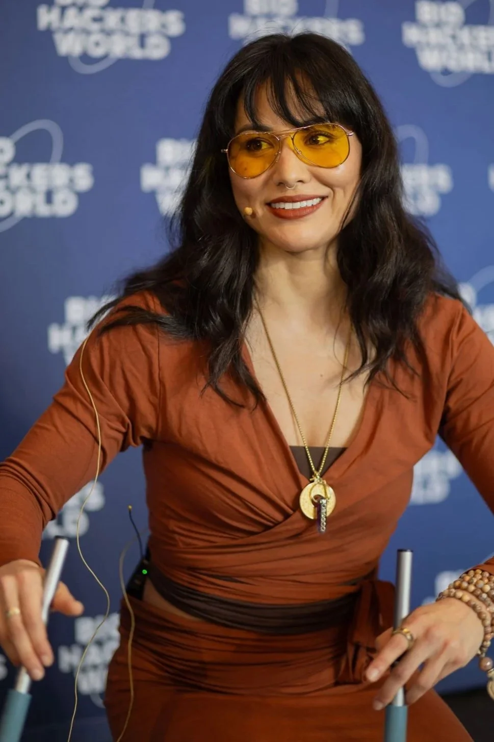 A woman with dark wavy hair and wearing yellow-tinted glasses, a burnt orange dress, and layered jewelry, smiling and sitting at a table with a blue background that has white text reading 'PIONEERS OF THE WORLD'.
