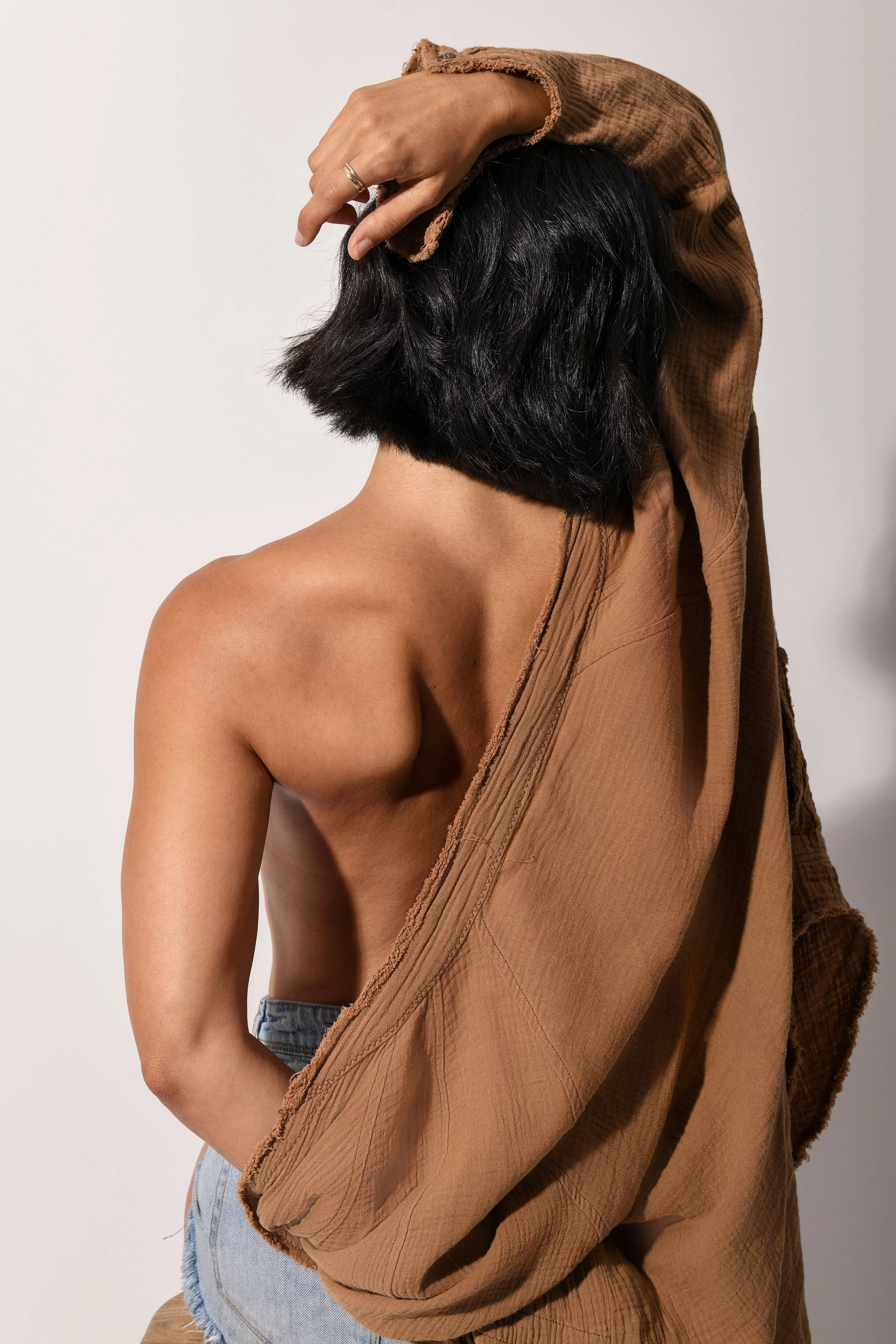 A woman with short black hair and nude skin, wearing a brown oversized jacket draped over her shoulder, and ripped denim shorts.