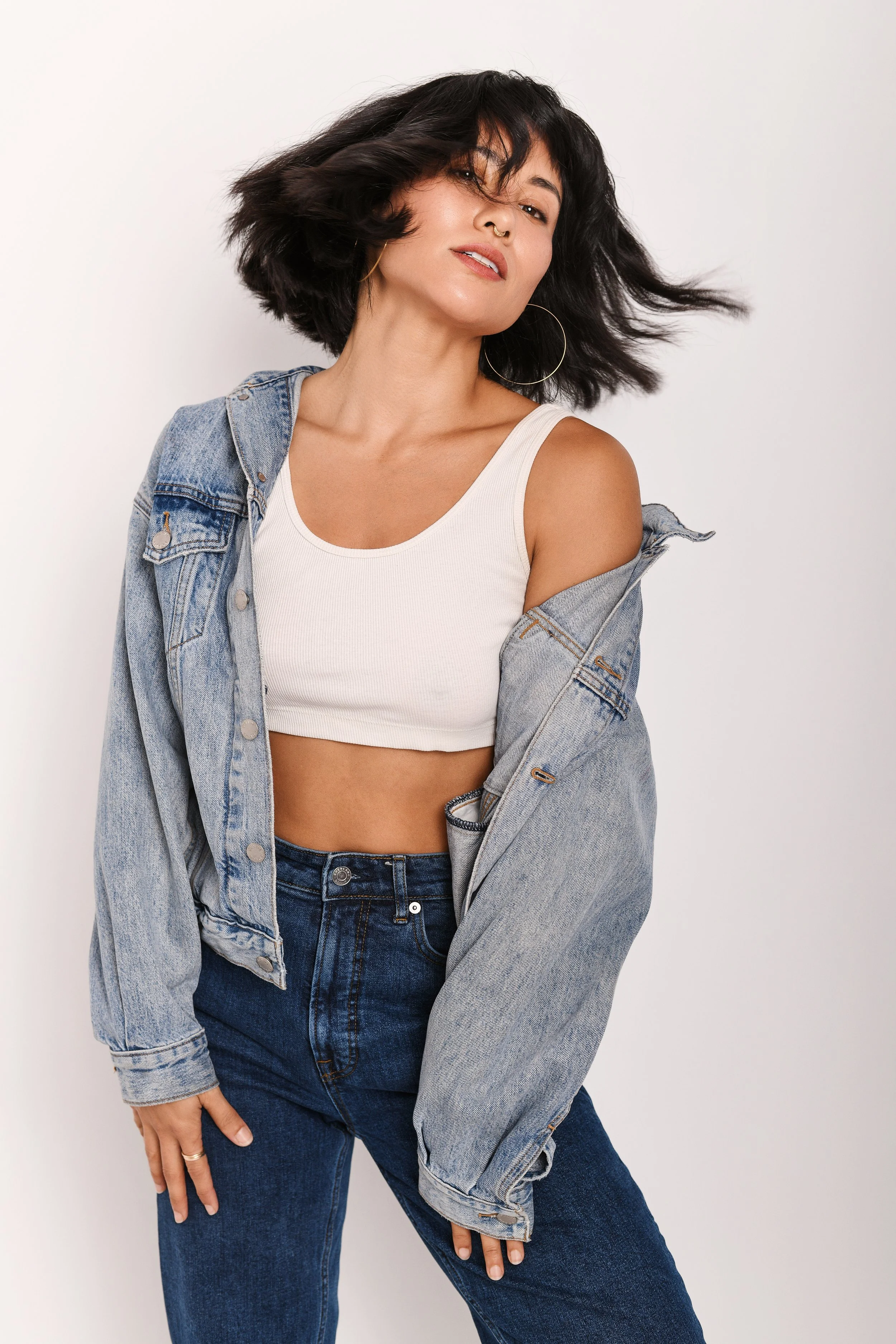 A woman with shoulder-length black hair wearing a white crop top, a light blue denim jacket off her shoulders, and dark blue jeans, posing against a plain white background.