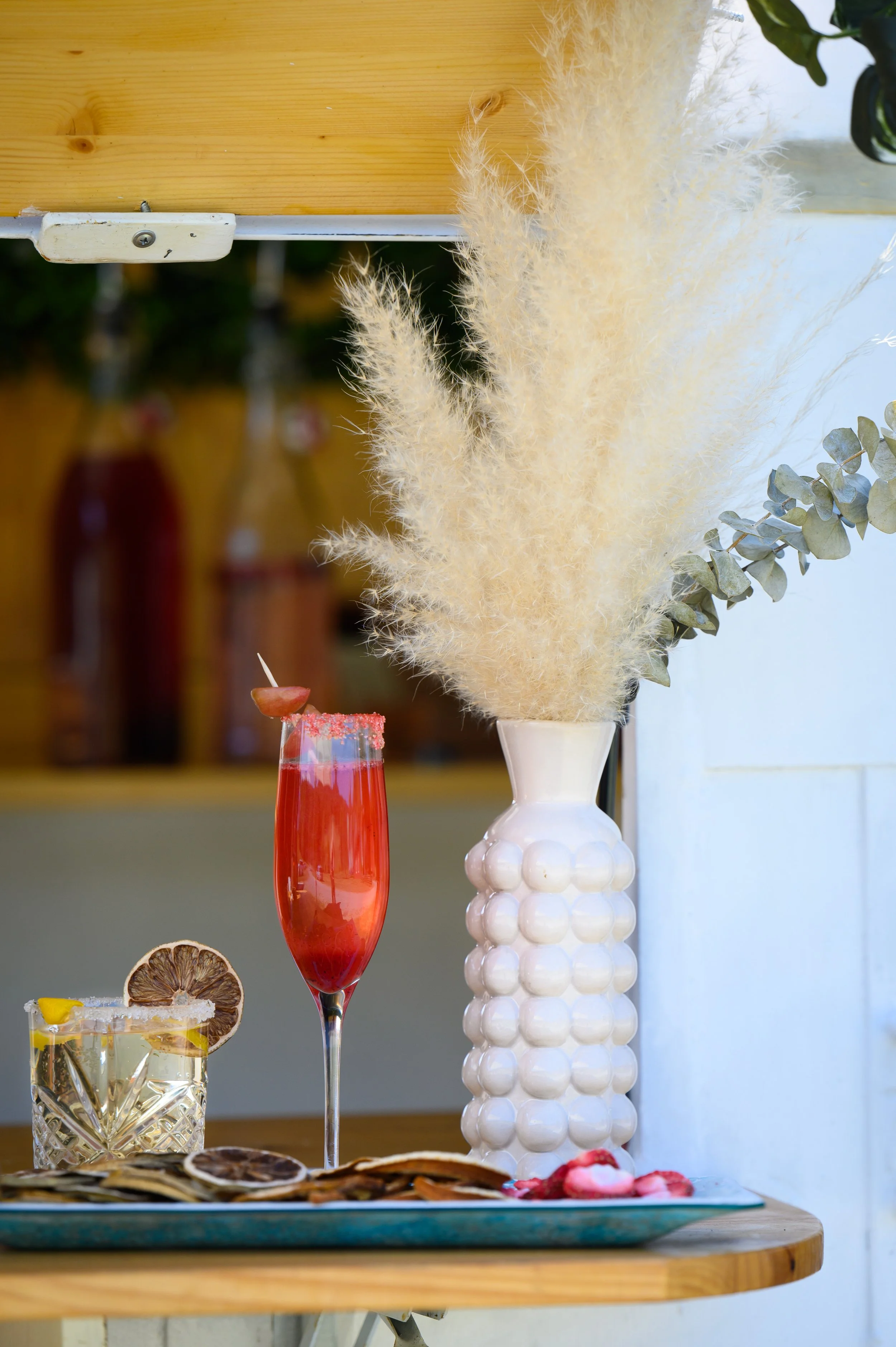Decorative arrangement of three cocktails on a wooden table, with a large white vase holding pampas grass and eucalyptus leaves in the background.