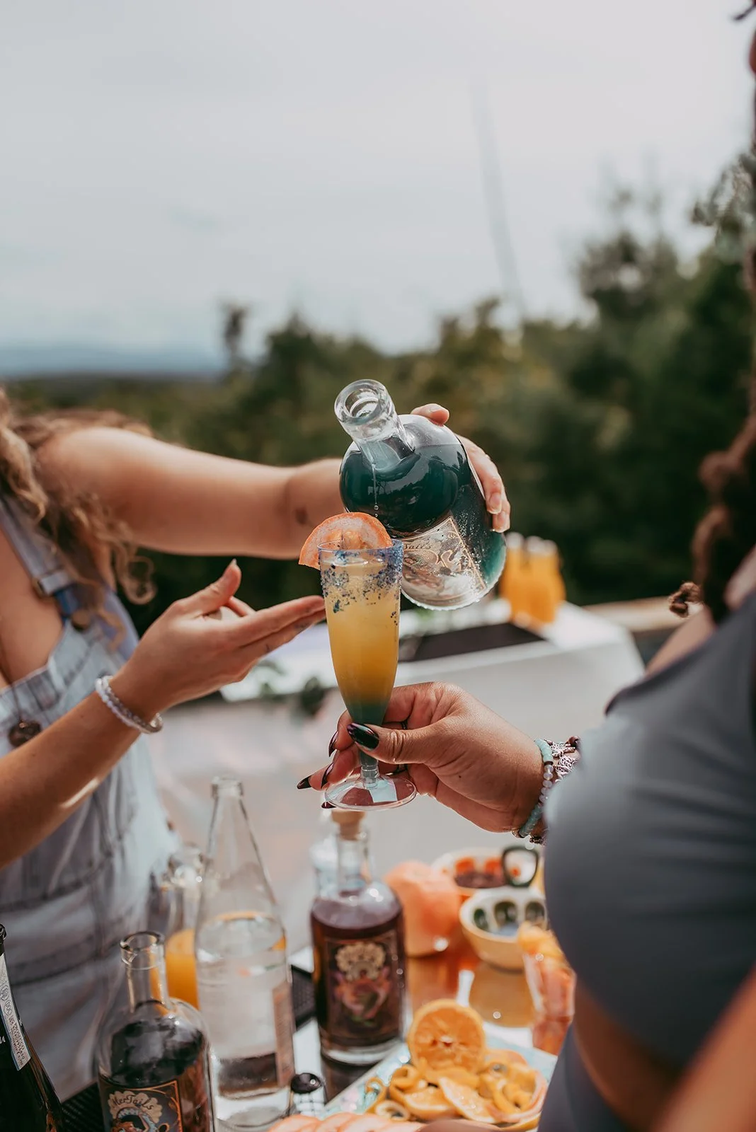 Two people sharing a toast with cocktails at an outdoor gathering, with a variety of drinks and snacks on the table.