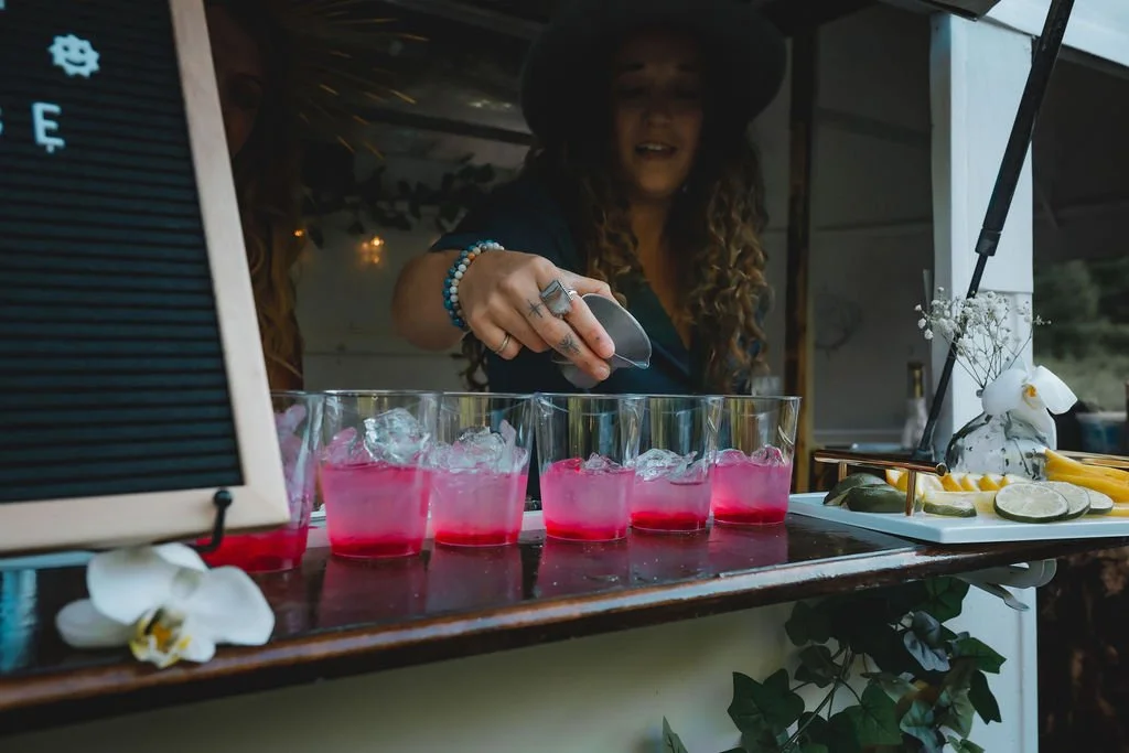 A woman in a hat adds liquid to pink drinks lined up on a bar, with a wooden counter decorated with orchids and fruits, and a black menu board on the side.