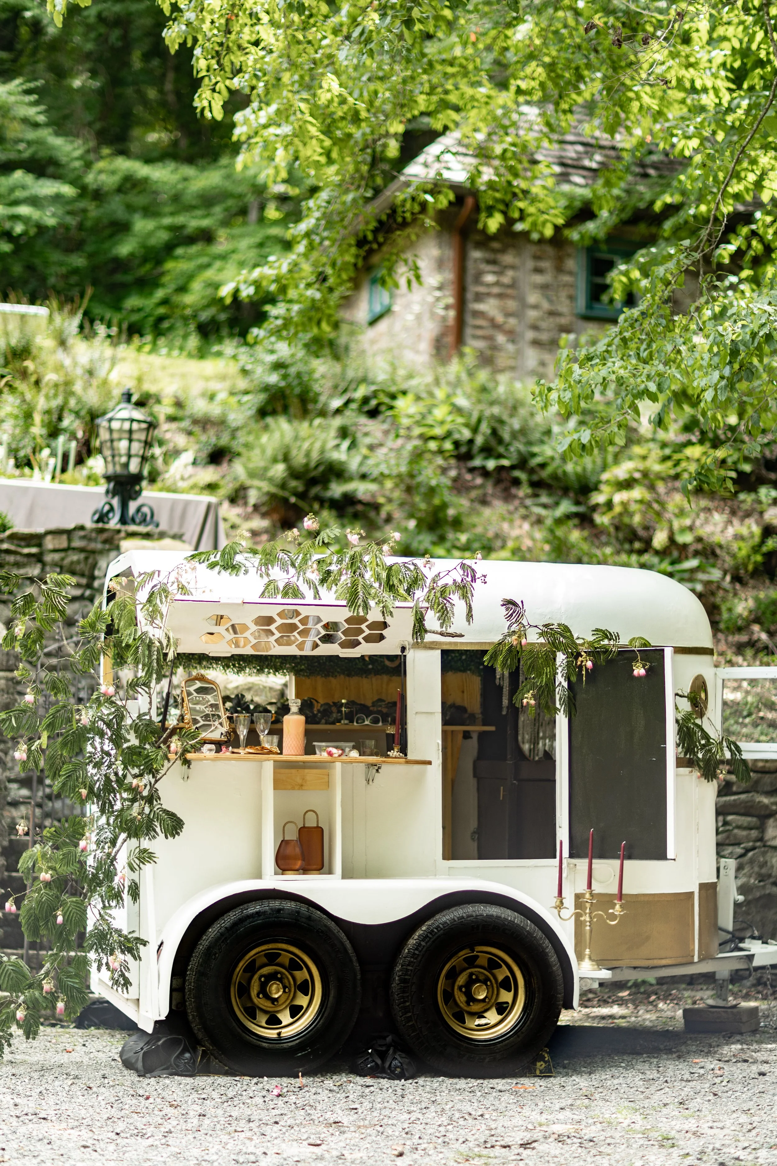 A vintage white food trailer decorated with greenery and pink flowers, set outdoors in a lush garden with a stone house and trees in the background.