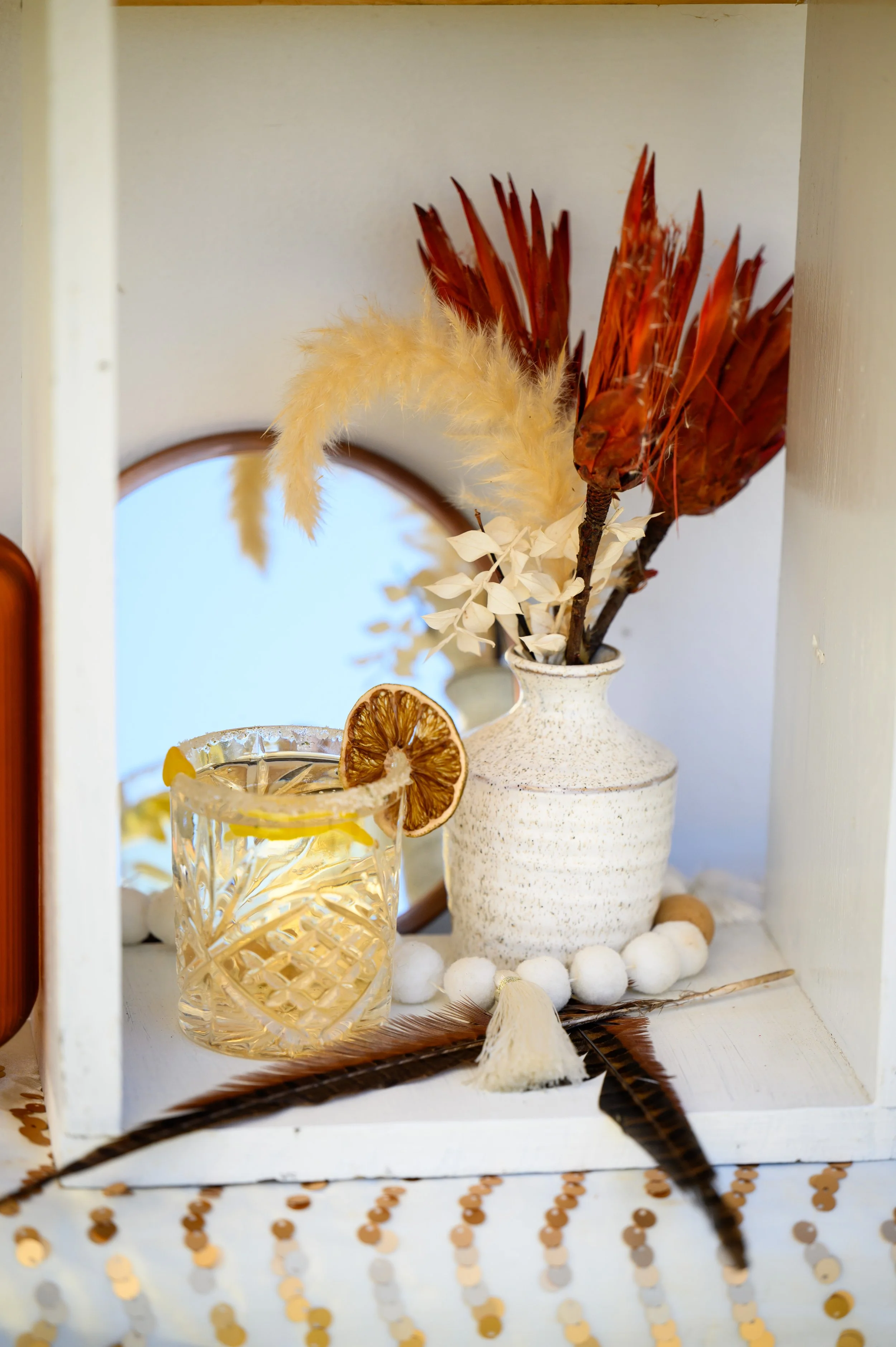 Decorative arrangement with a glass of lemon drink with dried lemon slice, a white vase with dried flowers and leaves, a feather, and white pom-poms on a white box, set against a white background with a mirror.