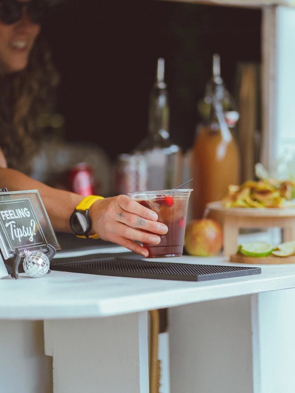 A person wearing a yellow watch holds a plastic cup of red drink with raspberries. On the counter is a small sign that says 'Feeling Tipsy?' along with a disco ball decoration. In the background, there are juice bottles, a wooden crate with sandwiches, and an apple near lime wedges, with a window letting in natural light.