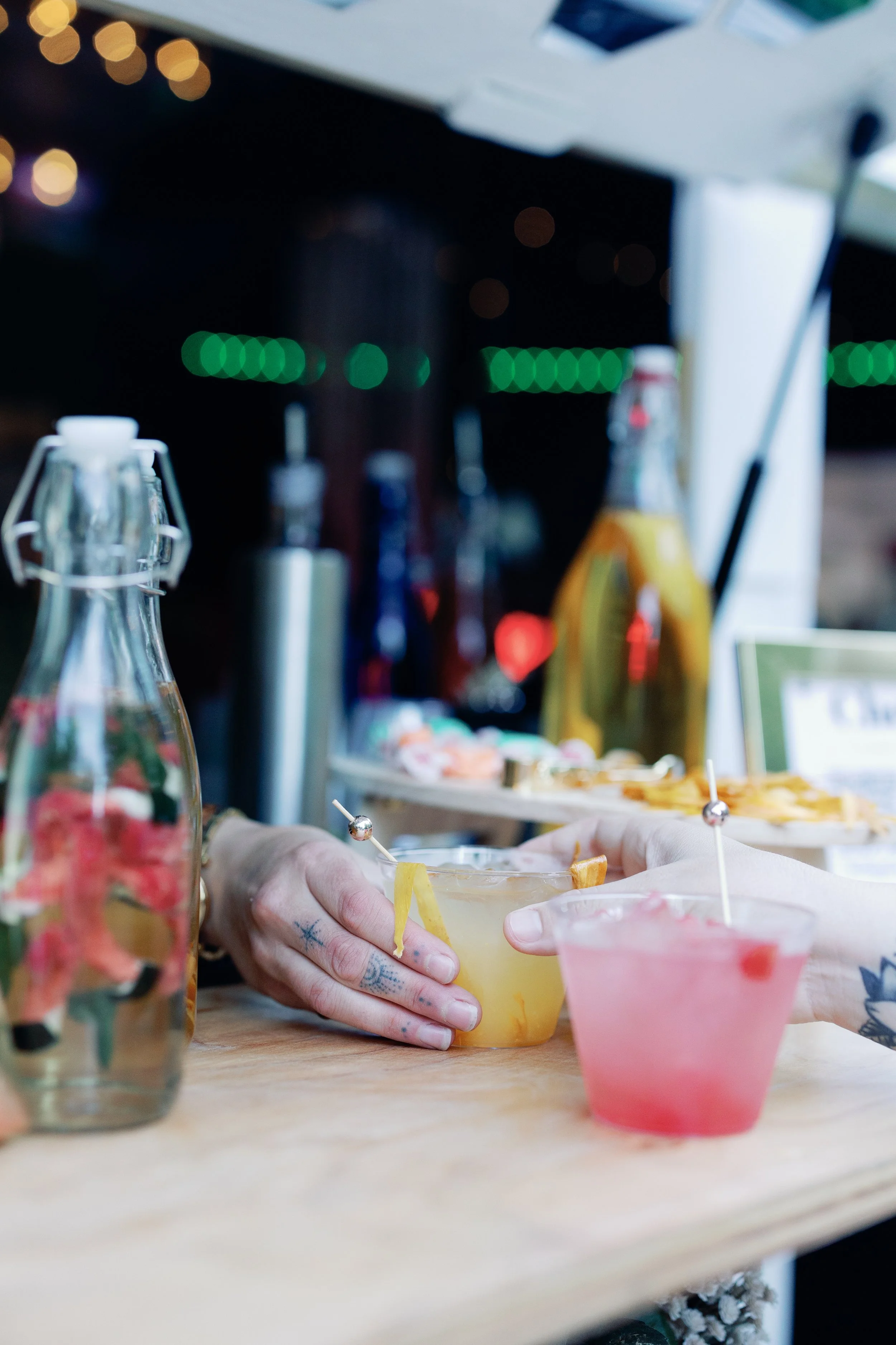 Two hands exchanging drinks at a bar, with various bottles and drinks in the background.
