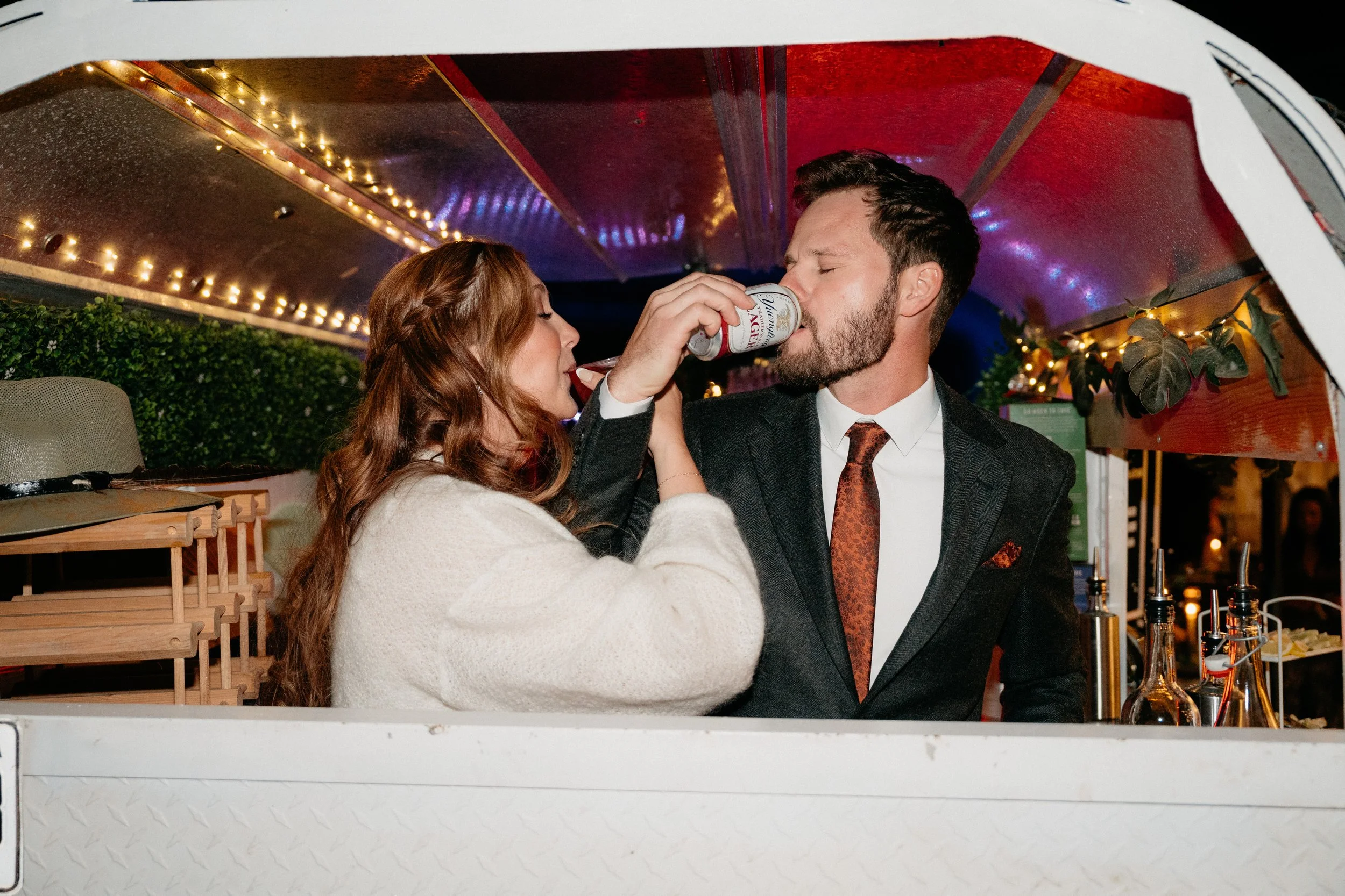 A woman and a man in formal attire sharing a beer inside a decorated food truck at night.