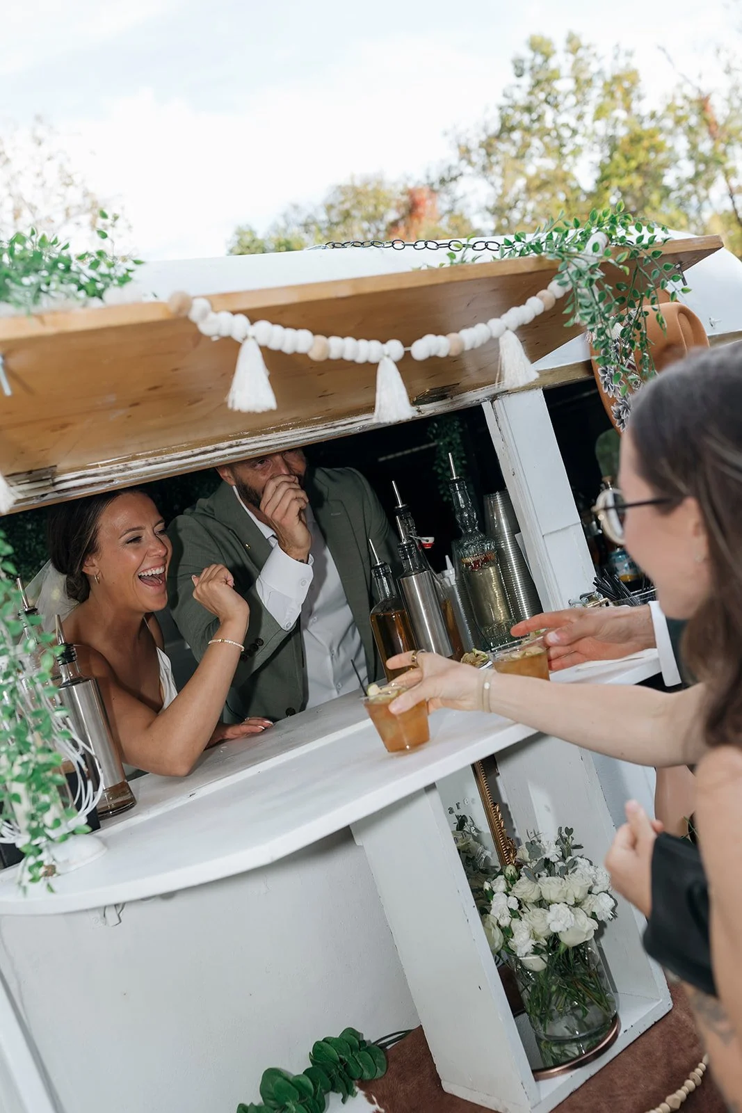 A bartender man serving drinks to two women at an outdoor bar, with a smiling woman in a white dress, a man in a suit, and a woman with glasses in the foreground. There are decorative plants and a floral arrangement below the bar.