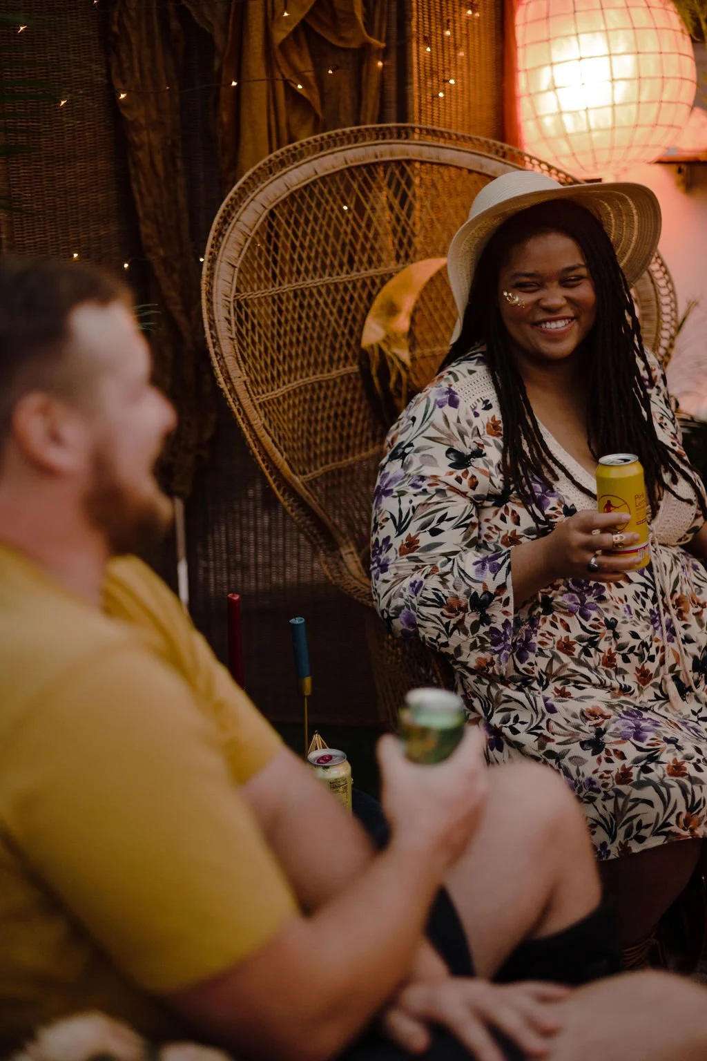 Two friends enjoying drinks at a cozy, dimly-lit gathering with decorative wicker wall art, fairy lights, and warm ambiance.