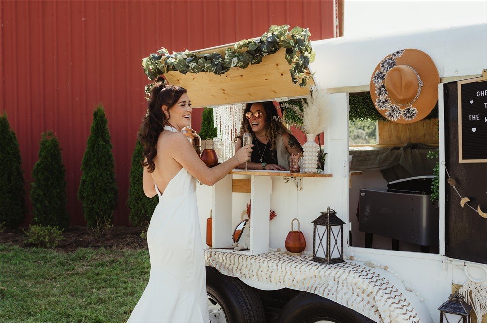 A woman in a white dress is enjoying a drink at a mobile beverage stand decorated with a green leafy garland. She is smiling and interacting with a woman behind the stand, who is wearing sunglasses and a hat. The stand has a rustic style with a red barn wall in the background and various decorative items including a hat, lanterns, and vases.