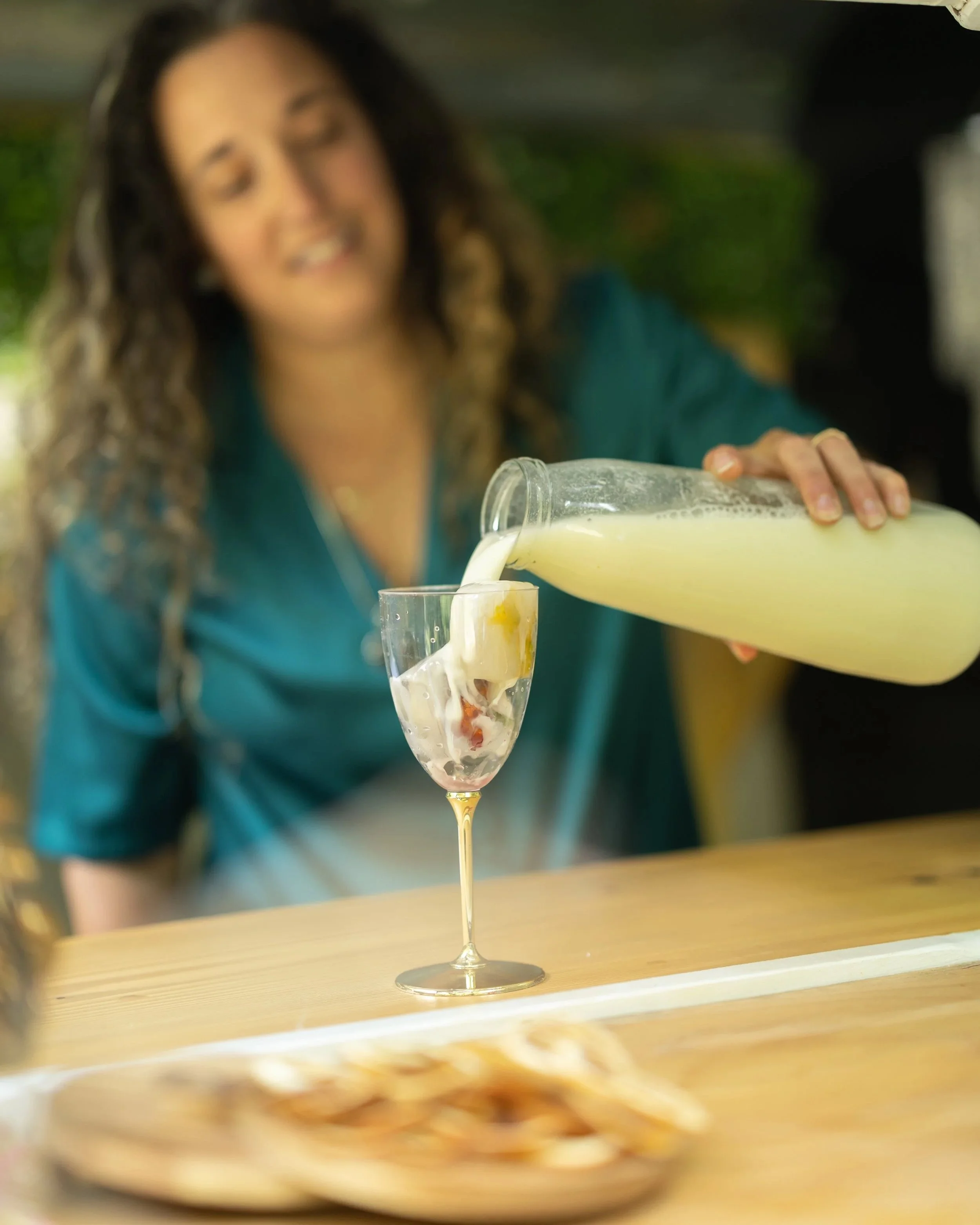 A woman pouring a yellow beverage from a glass pitcher into a wine glass with fruit pieces inside, on a wooden table.