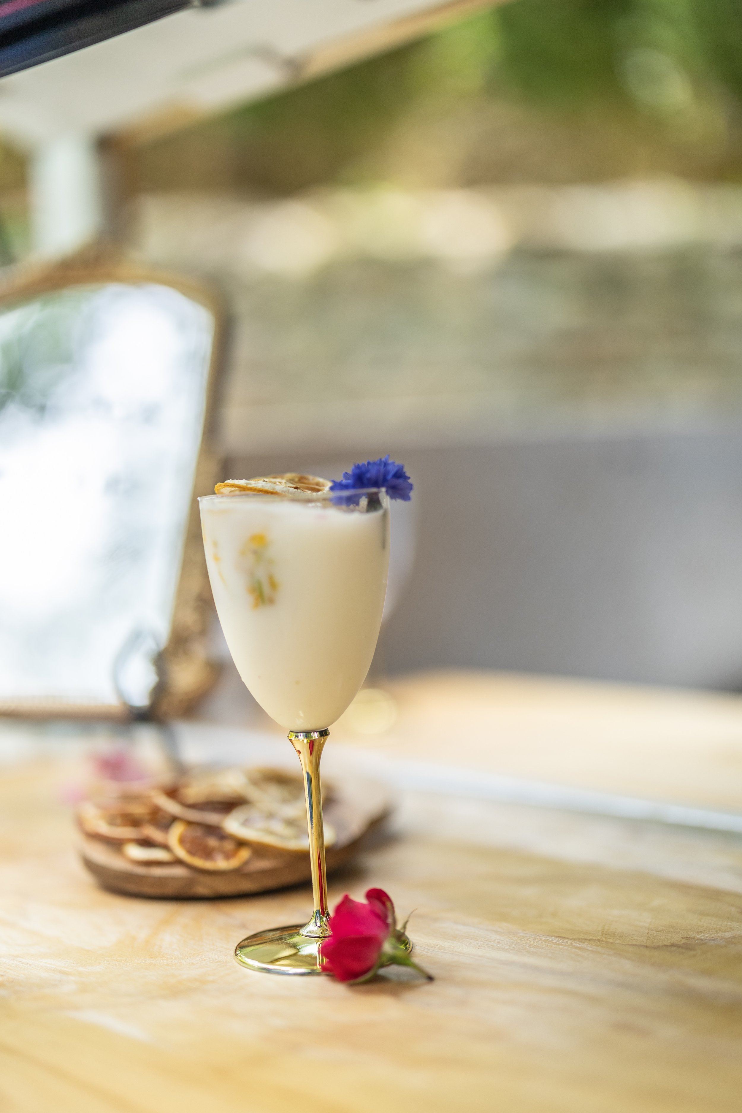 A glass of white cocktail garnished with a blue flower and a slice of lemon, placed on a wooden surface with a pink flower nearby, with a mirror and dried citrus slices in the background.