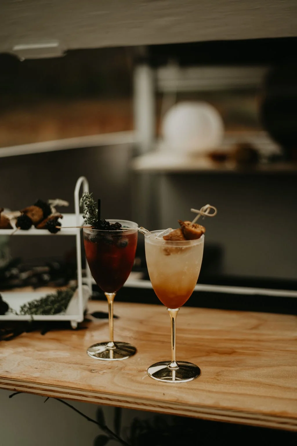Two elegant cocktails placed on a wooden surface with a starter tray of skewered food in the background.