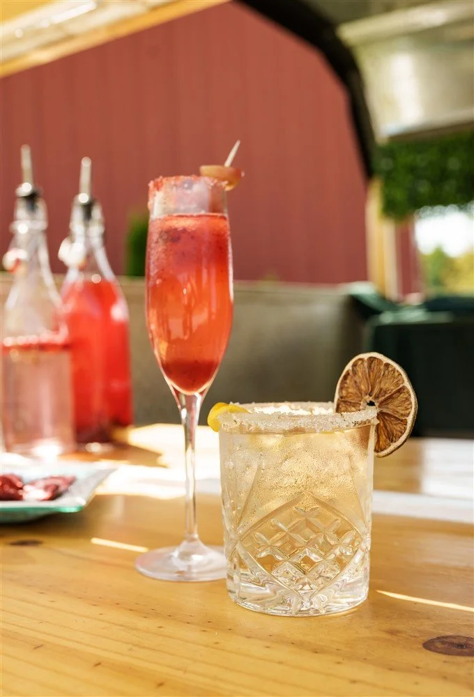A glass of margarita with a salted rim and a lemon wedge, and a tall glass of strawberry cocktail with a slice of orange, on a wooden table outside.