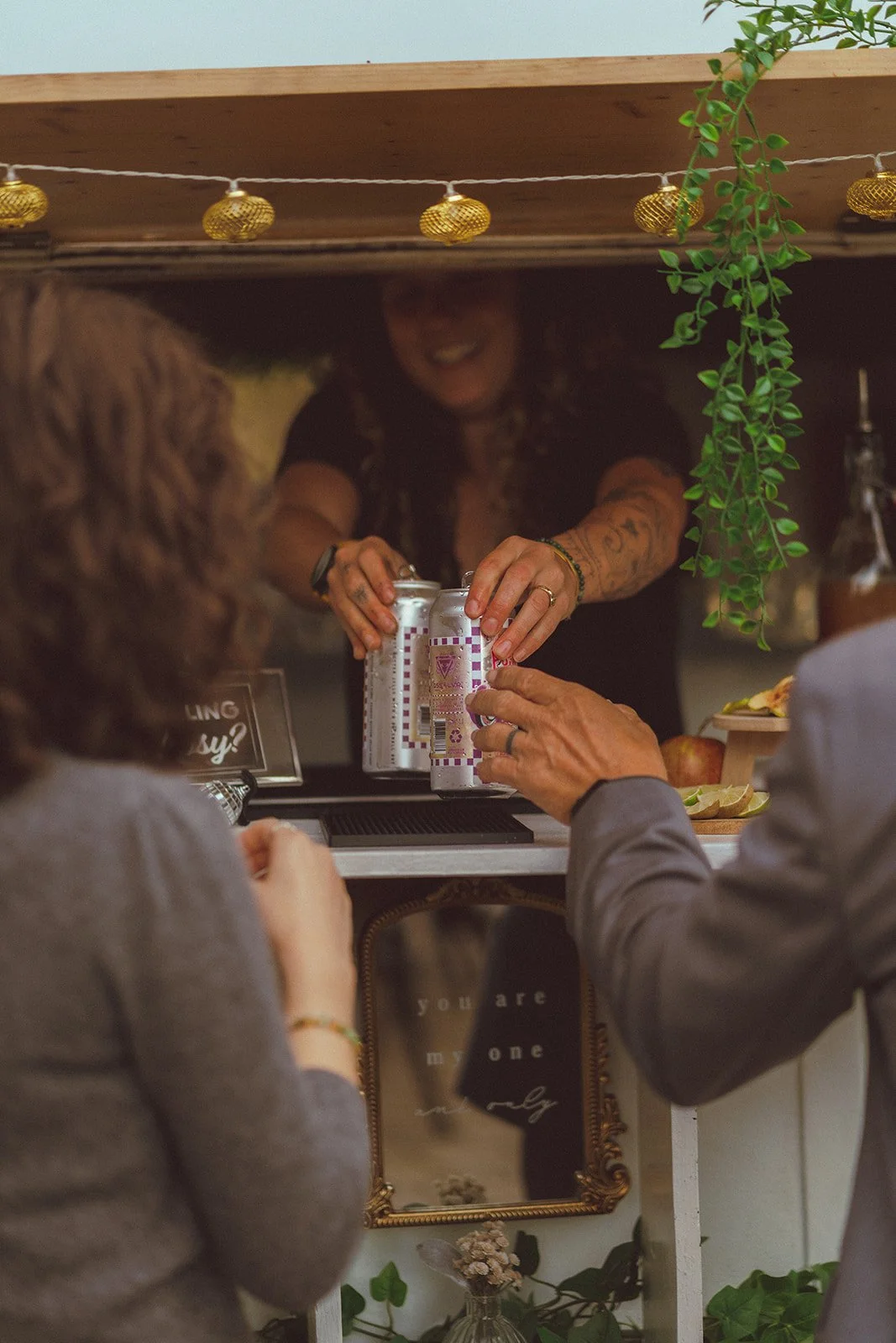 A person behind a bar is handing a drink to a customer, with a dog and another customer visible in the foreground, decorative string lights and green plants overhead, and a mirror with the words 'you are my one' reflected below.