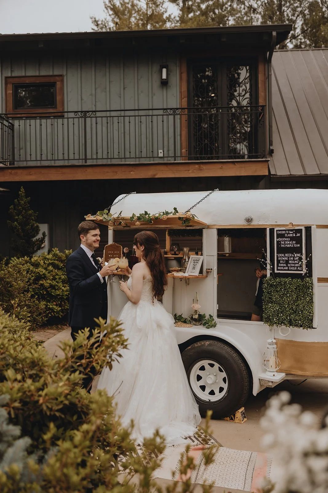 Bride and groom sharing drinks in front of a white vintage trailer at an outdoor wedding celebration.
