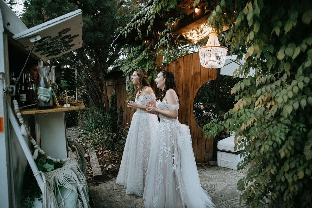 Two women in white wedding dresses having a conversation outdoors near a wooden fence and lush greenery at dusk, with string lights and a chandelier hanging overhead.