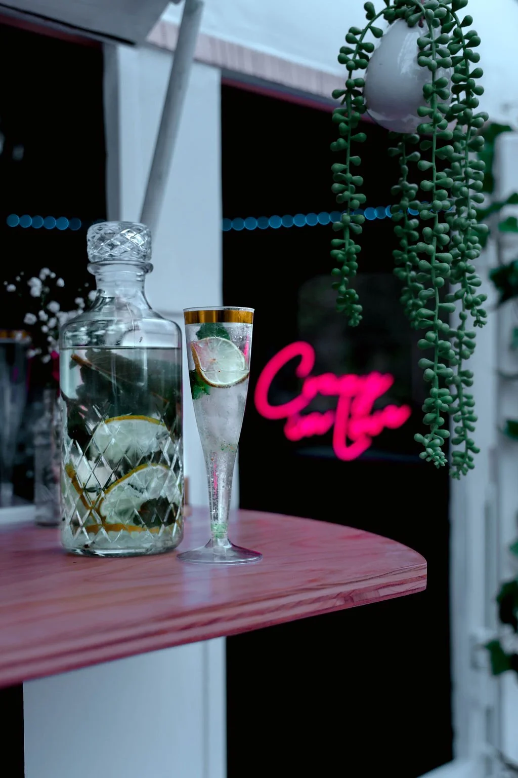 A glass pitcher of water with lemon slices and a glass of water with lemon slices on a pink wooden table, with a neon pink sign in the background and hanging green plants.